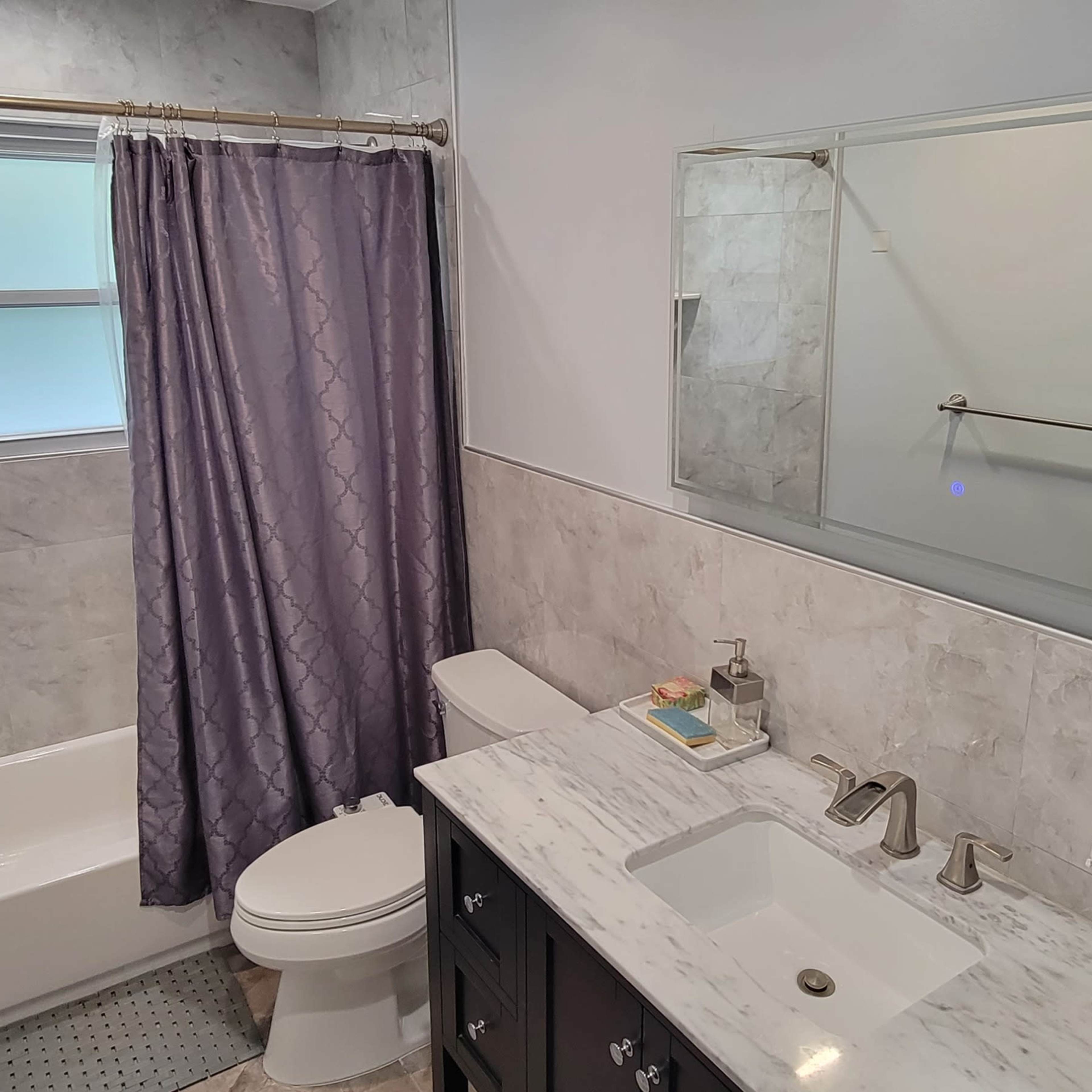 A modern bathroom featuring a shower-tub combination, a vanity with a marble countertop, and a window covered by a purple curtain.