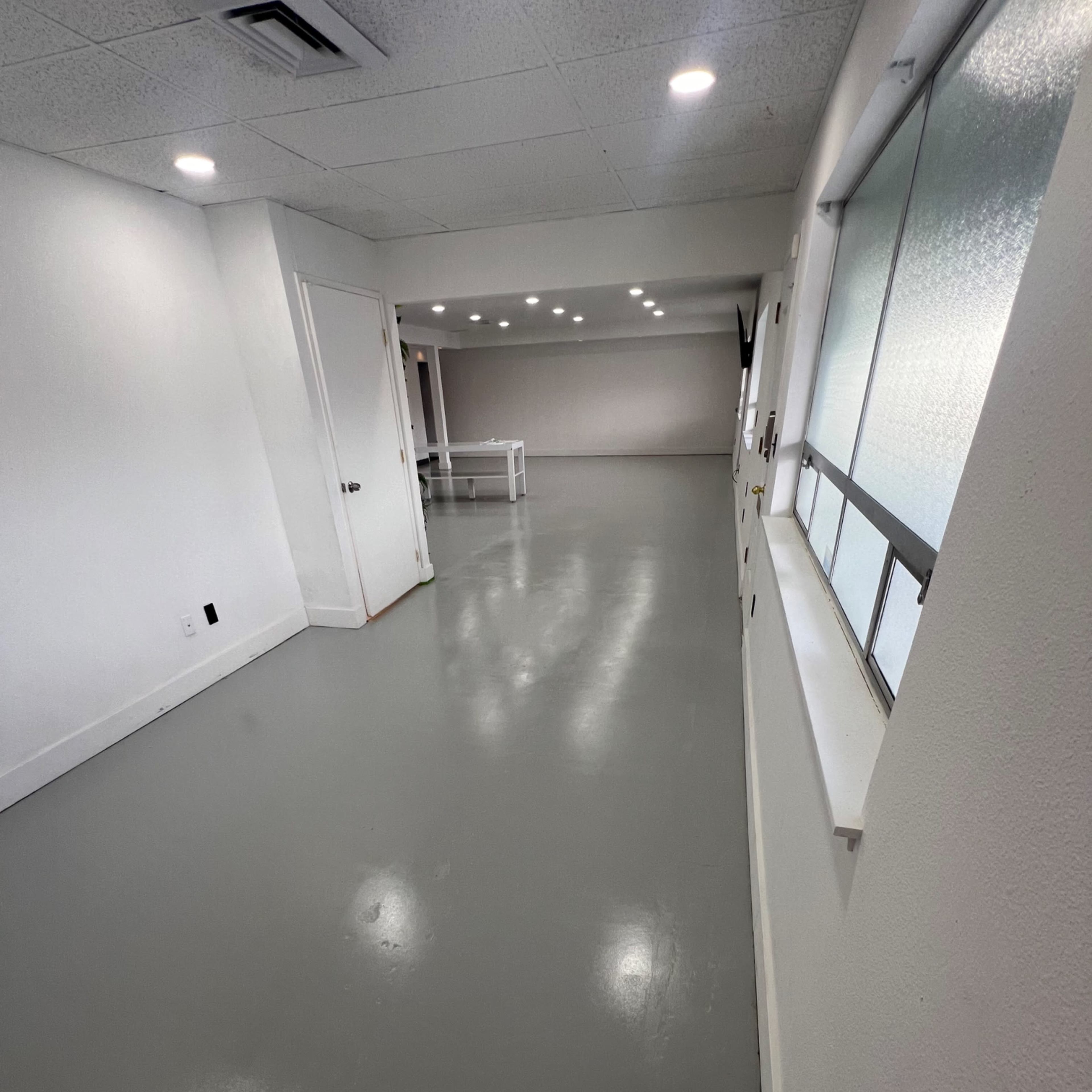 A spacious indoor area with gray flooring, white walls, and multiple lights overhead, leading to another room.