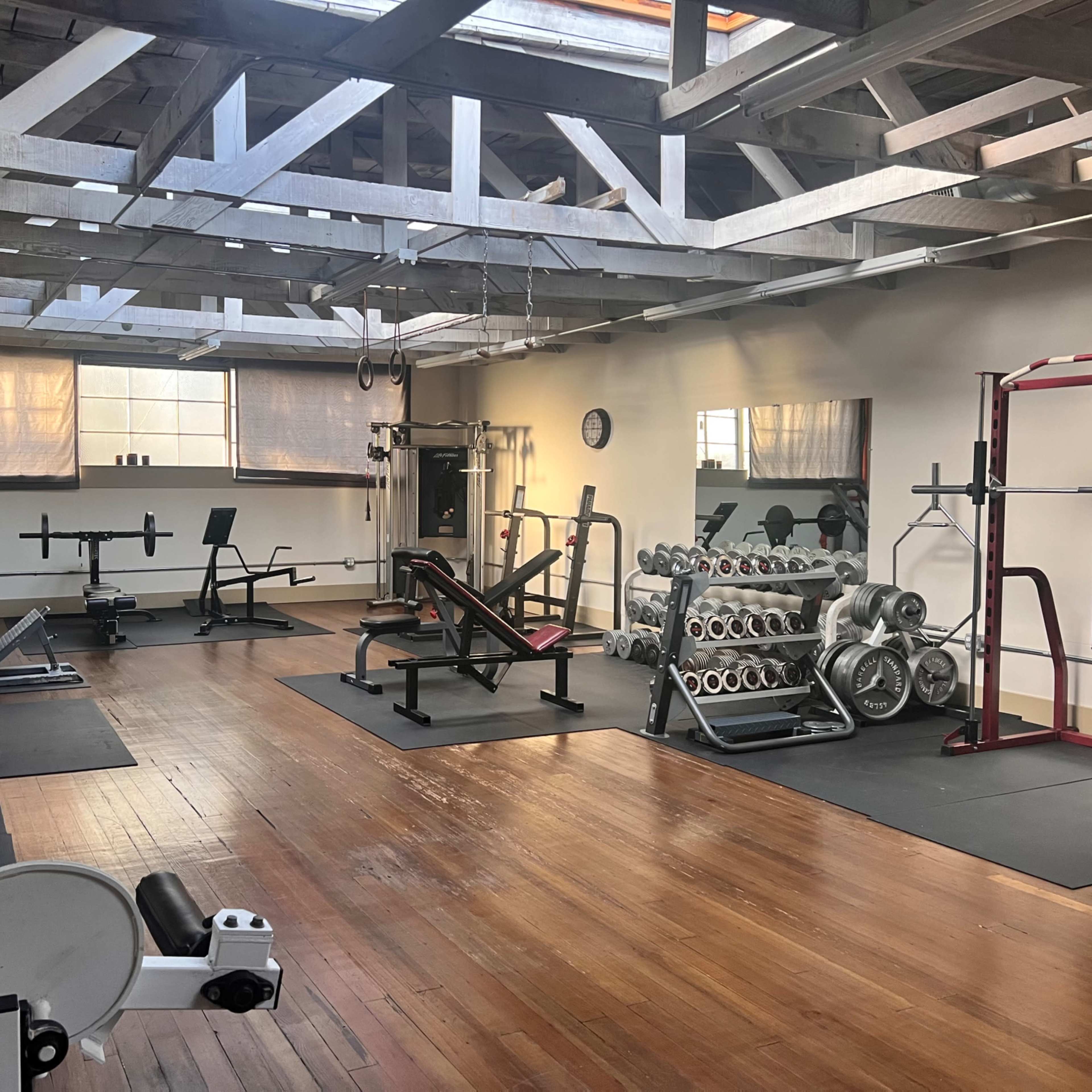 A spacious gym with various weightlifting equipment, benches, and racks, all arranged neatly on a wooden floor under a skylighted ceiling.