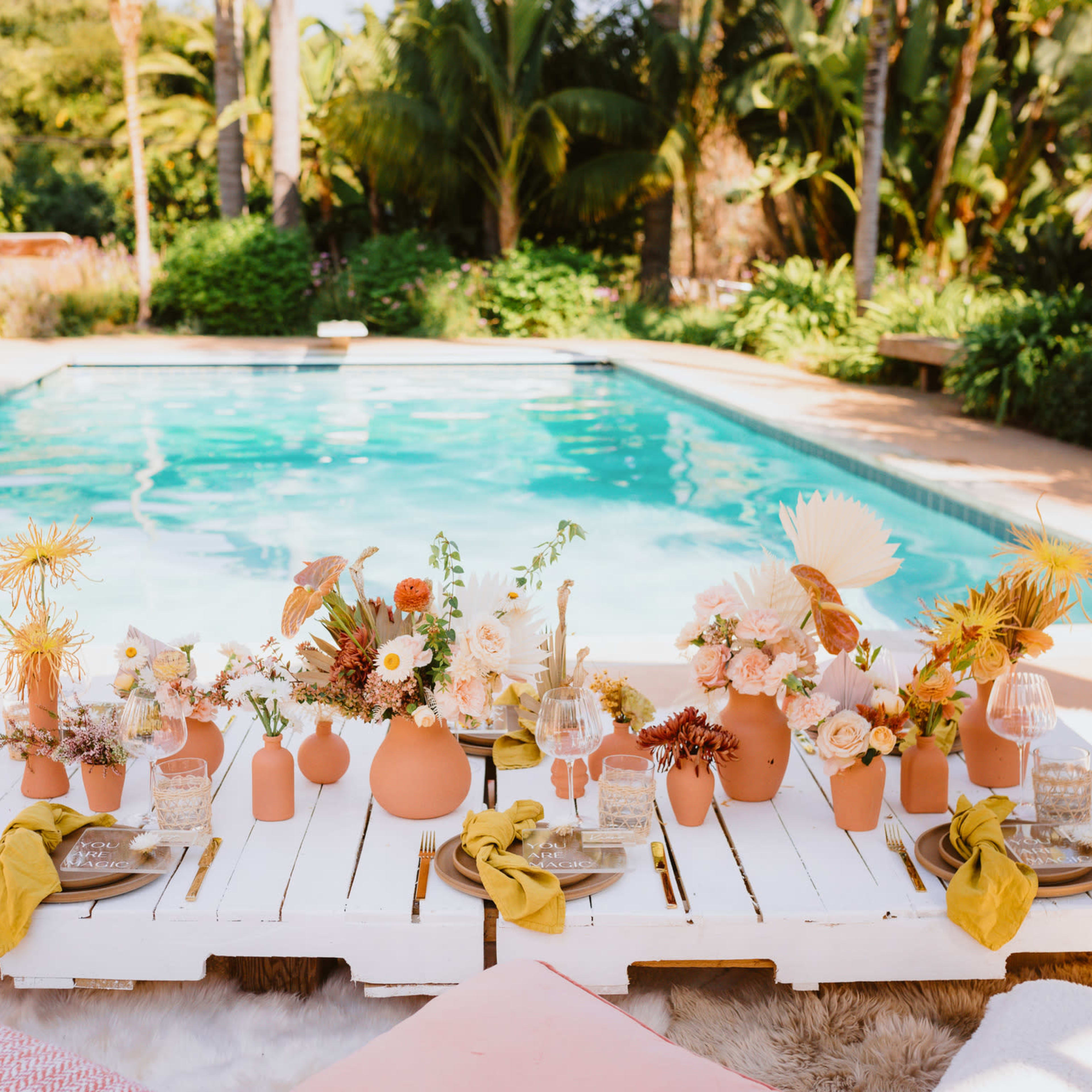 A styled outdoor dining setup featuring a wooden table adorned with colorful floral arrangements and tableware beside a swimming pool.