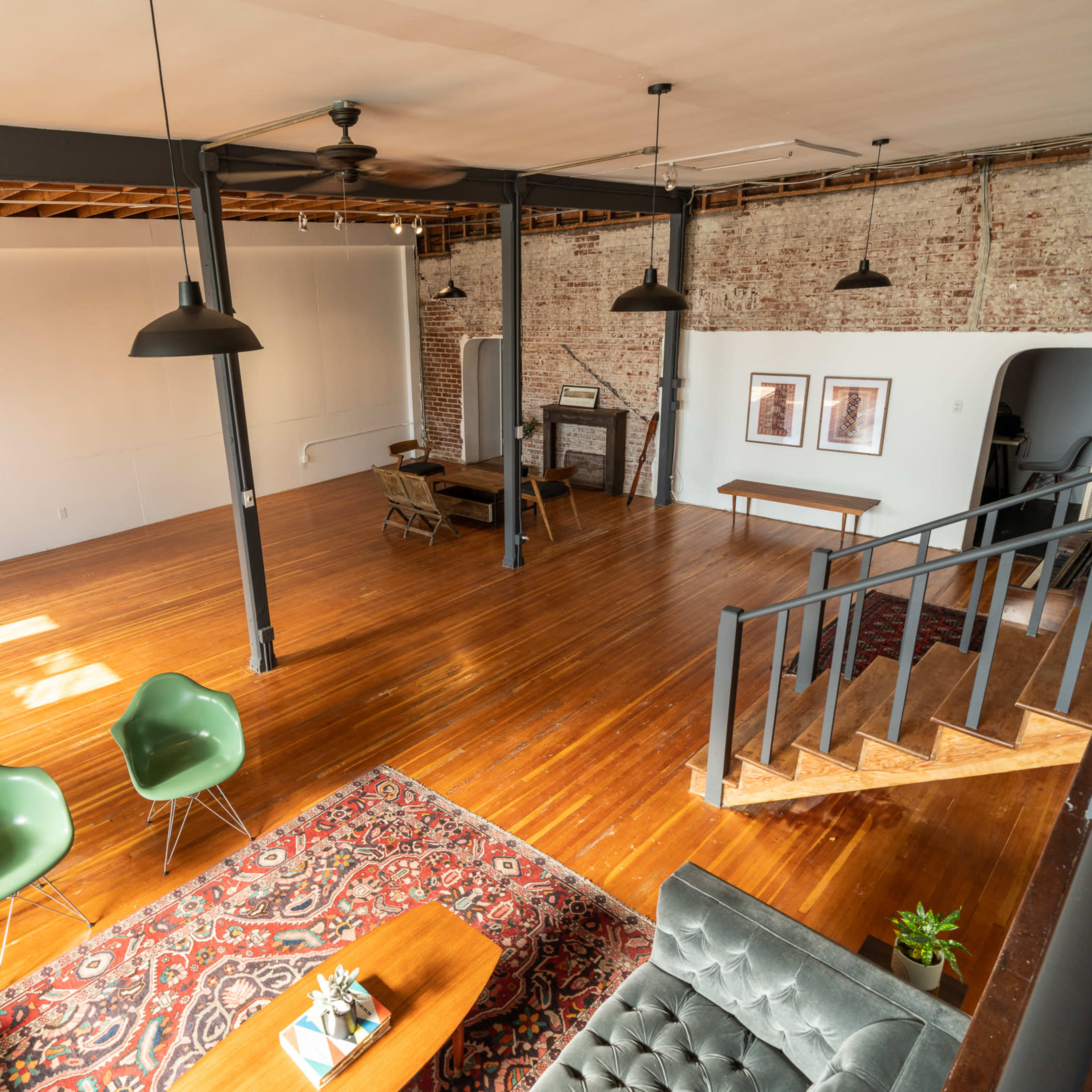 A spacious, open loft-style room with wooden floors, exposed brick walls, and modern furnishings, including green chairs and a gray sofa.