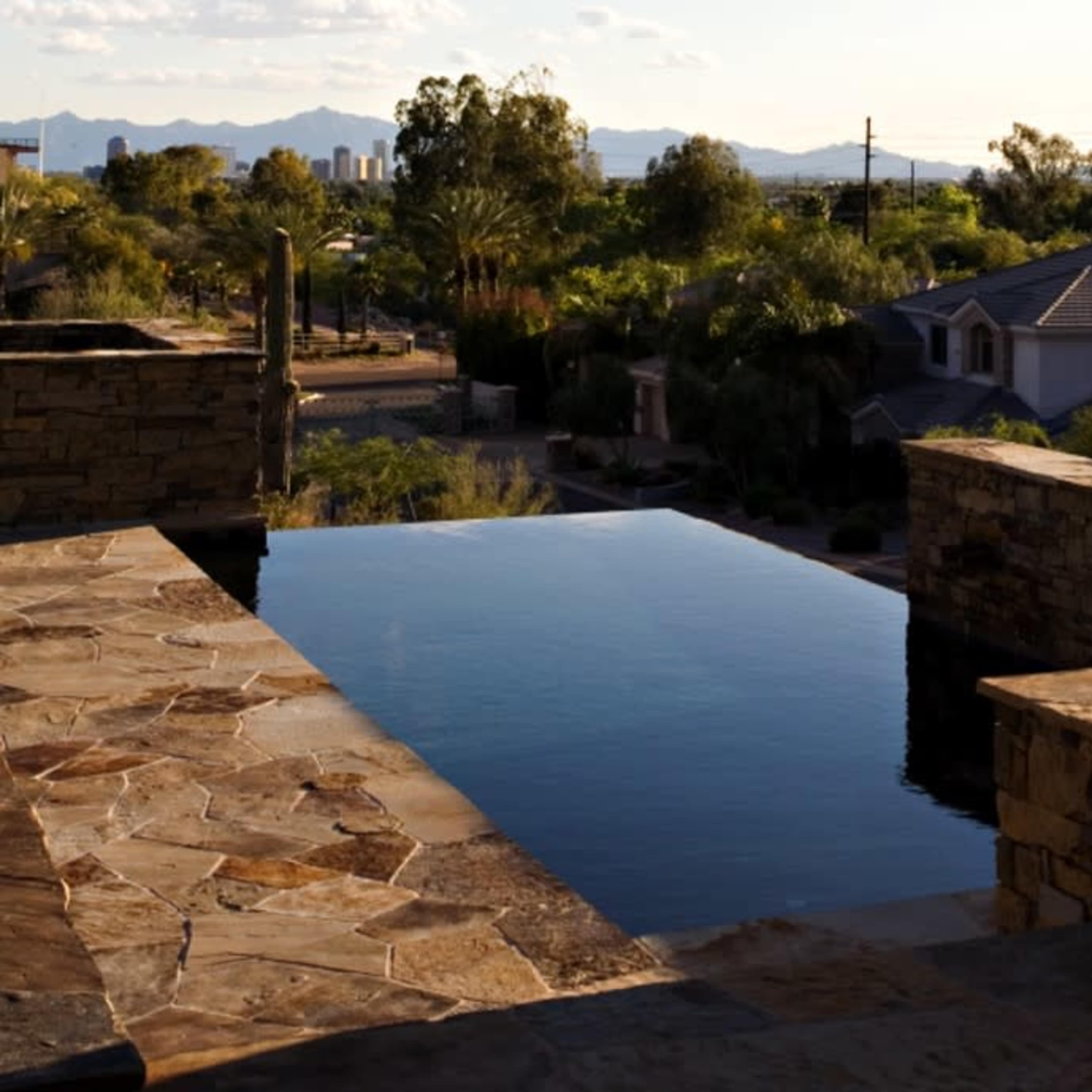 A stone deck surrounding an infinity pool with a view of distant mountains and a city skyline under a clear sky.