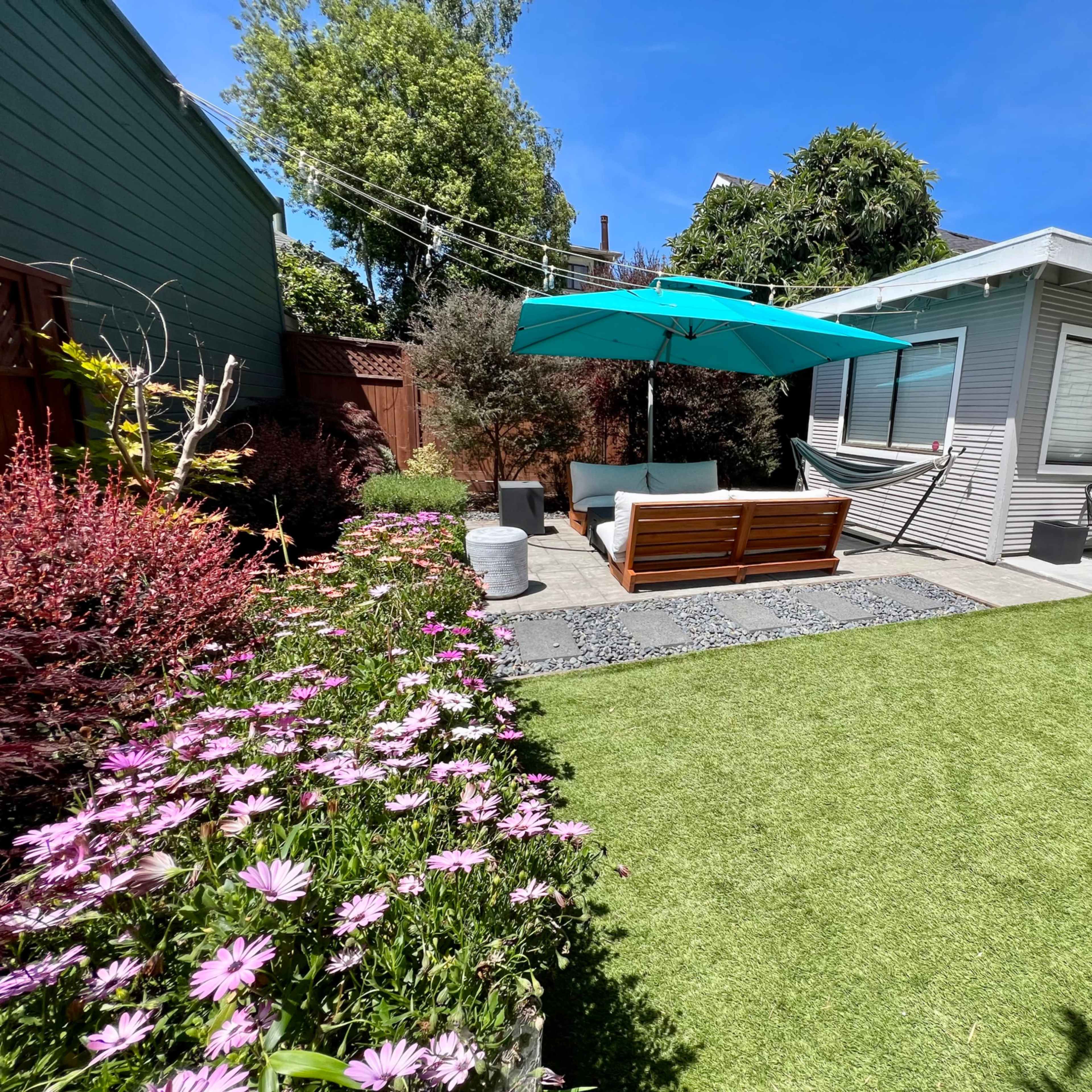 A landscaped backyard featuring a seating area under a blue umbrella, surrounded by flowers and artificial grass.