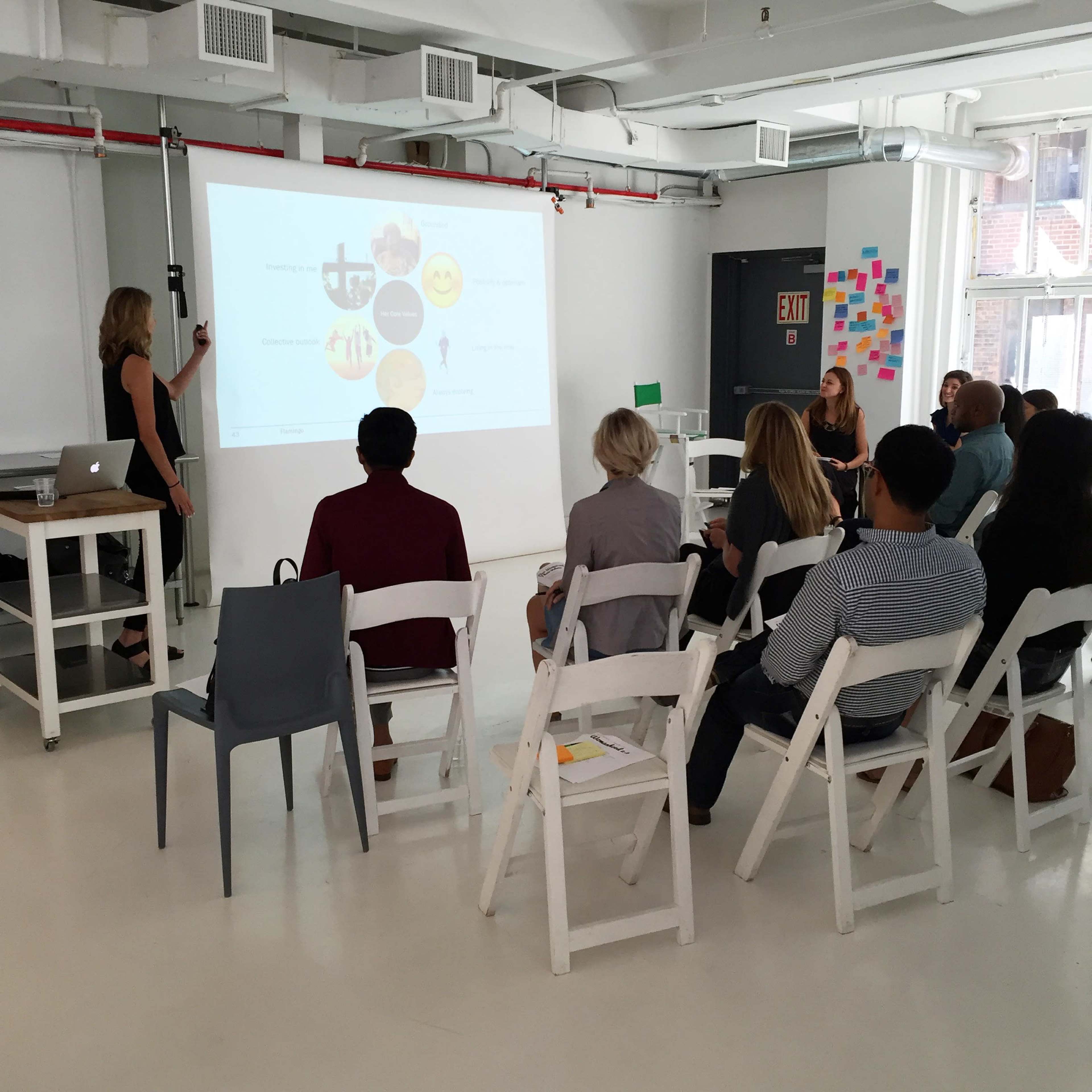 A presenter stands at the front of a modern room, displaying a visual presentation to an audience seated in white chairs.