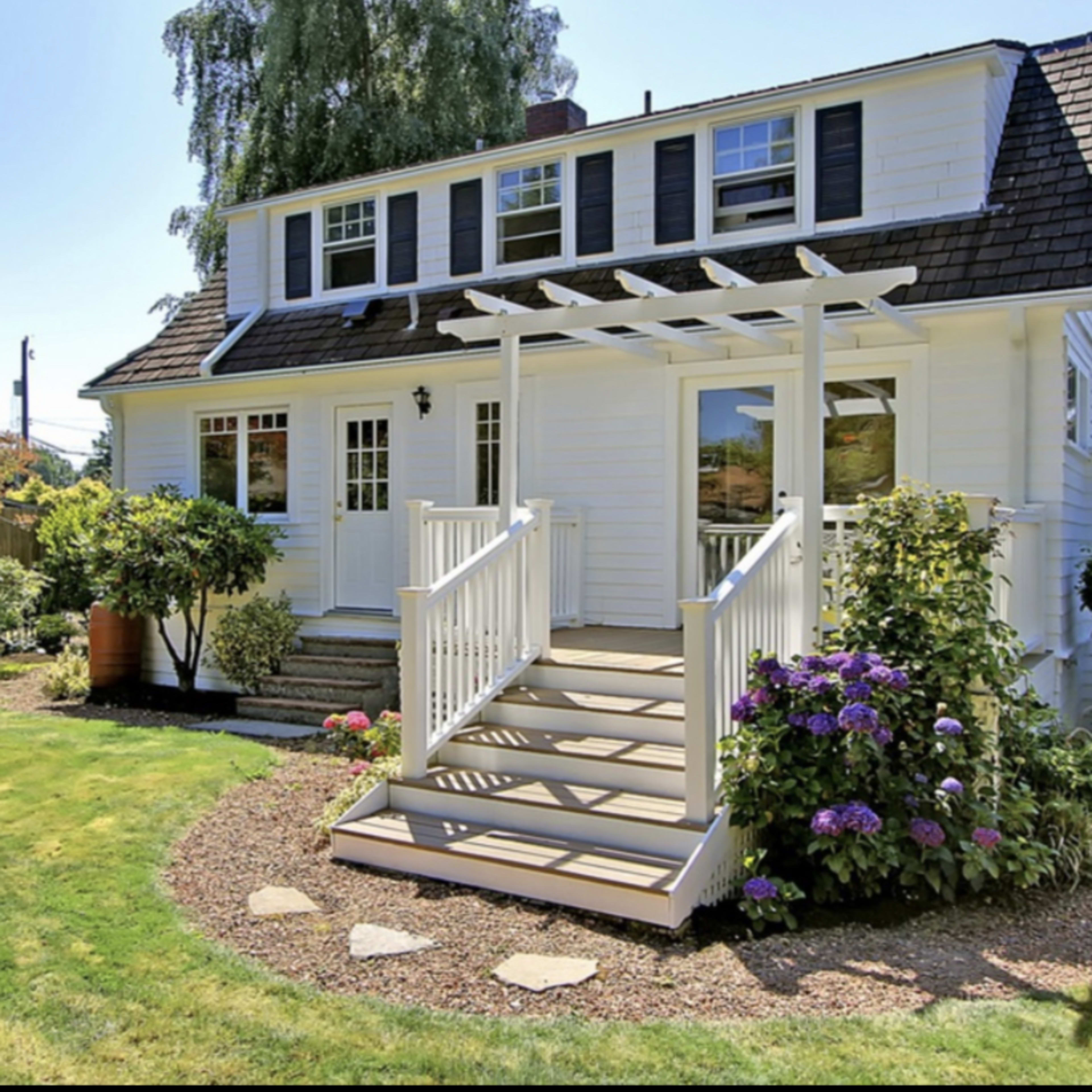 A two-story white house with a gabled roof features a front porch with a railing and landscaped gardens.