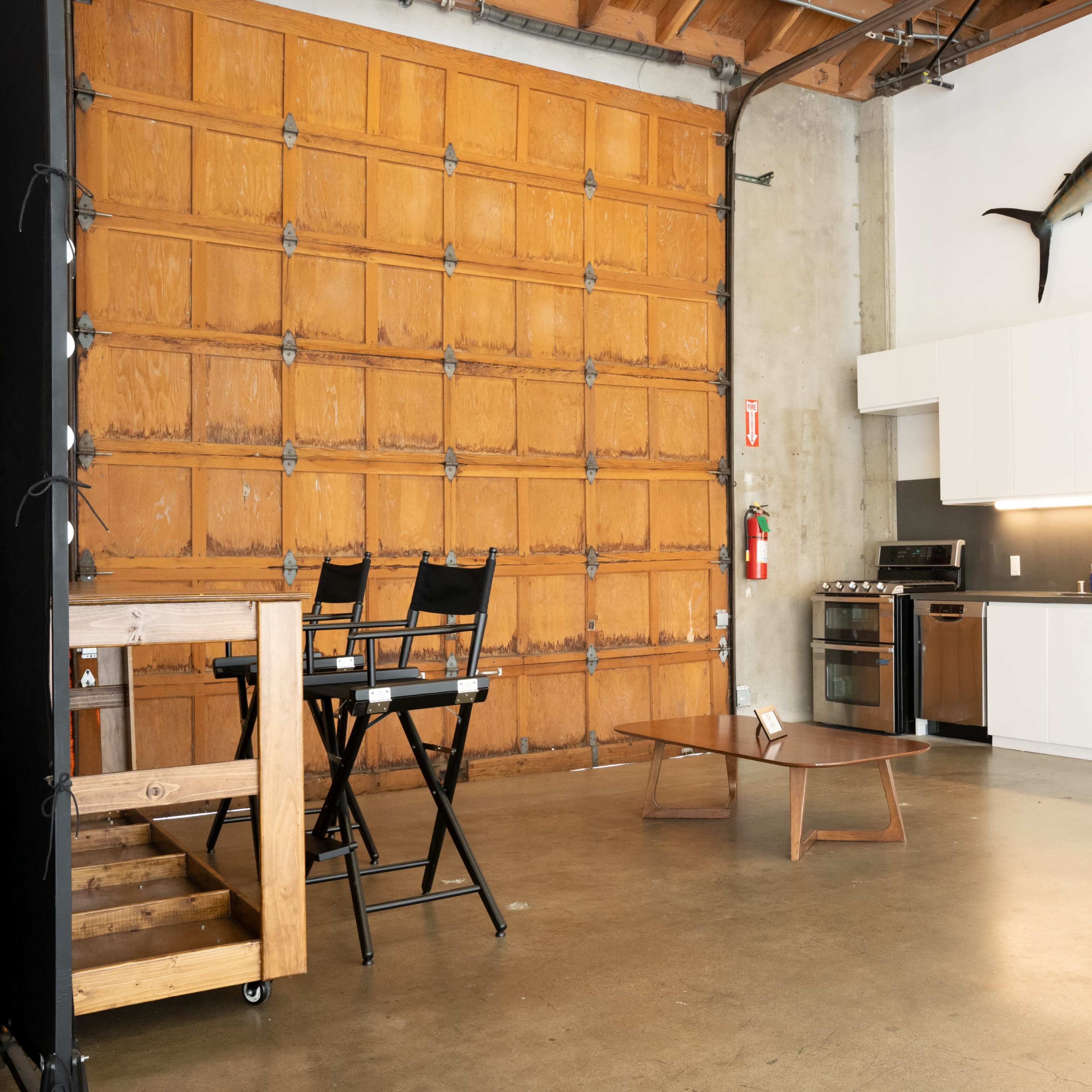 A spacious industrial-style kitchen and living area with a wooden garage door, a mounted fish above a modern kitchen setup, and minimal furniture.