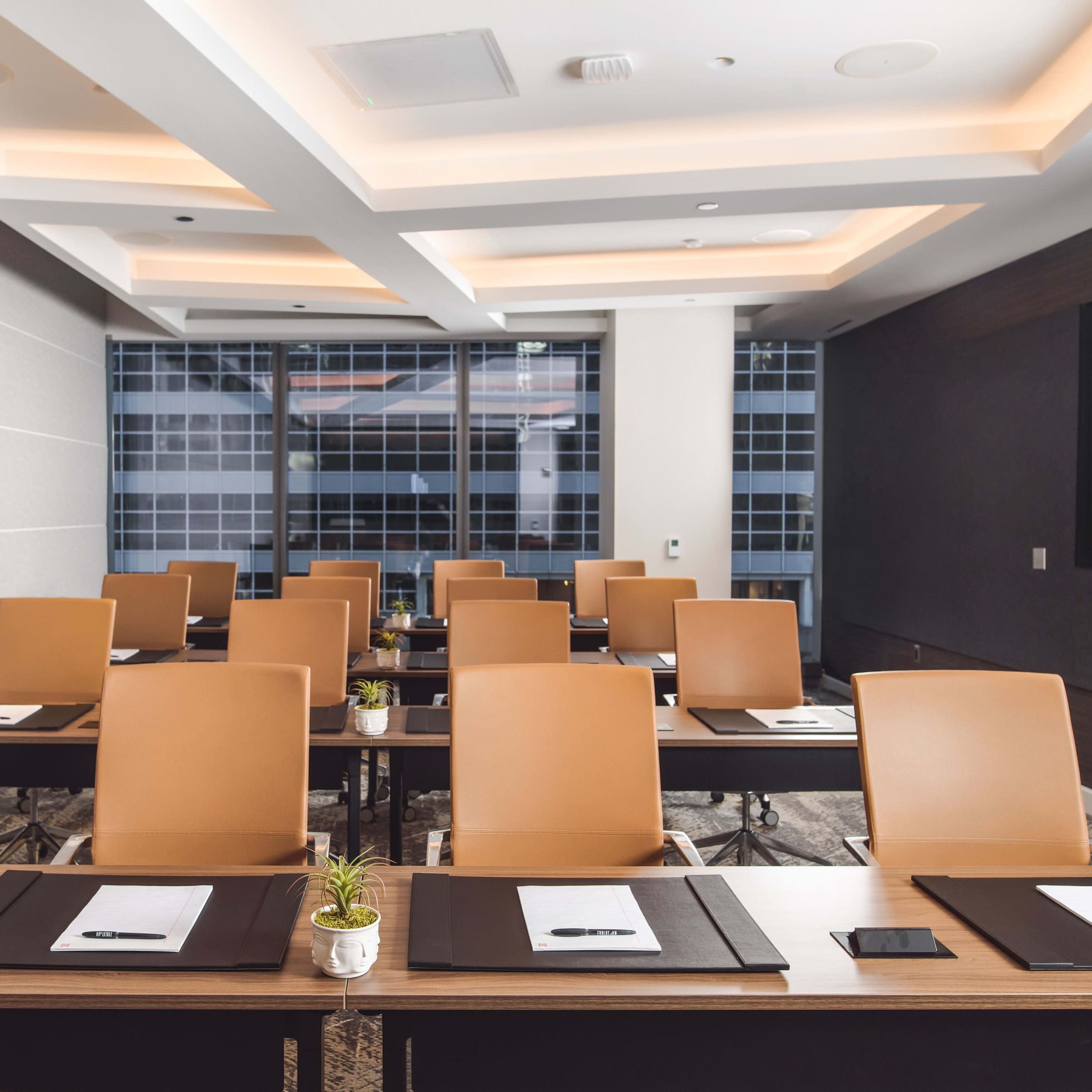 A modern conference room features several rows of brown chairs at wooden tables, with notebooks and small plants placed at each setting.