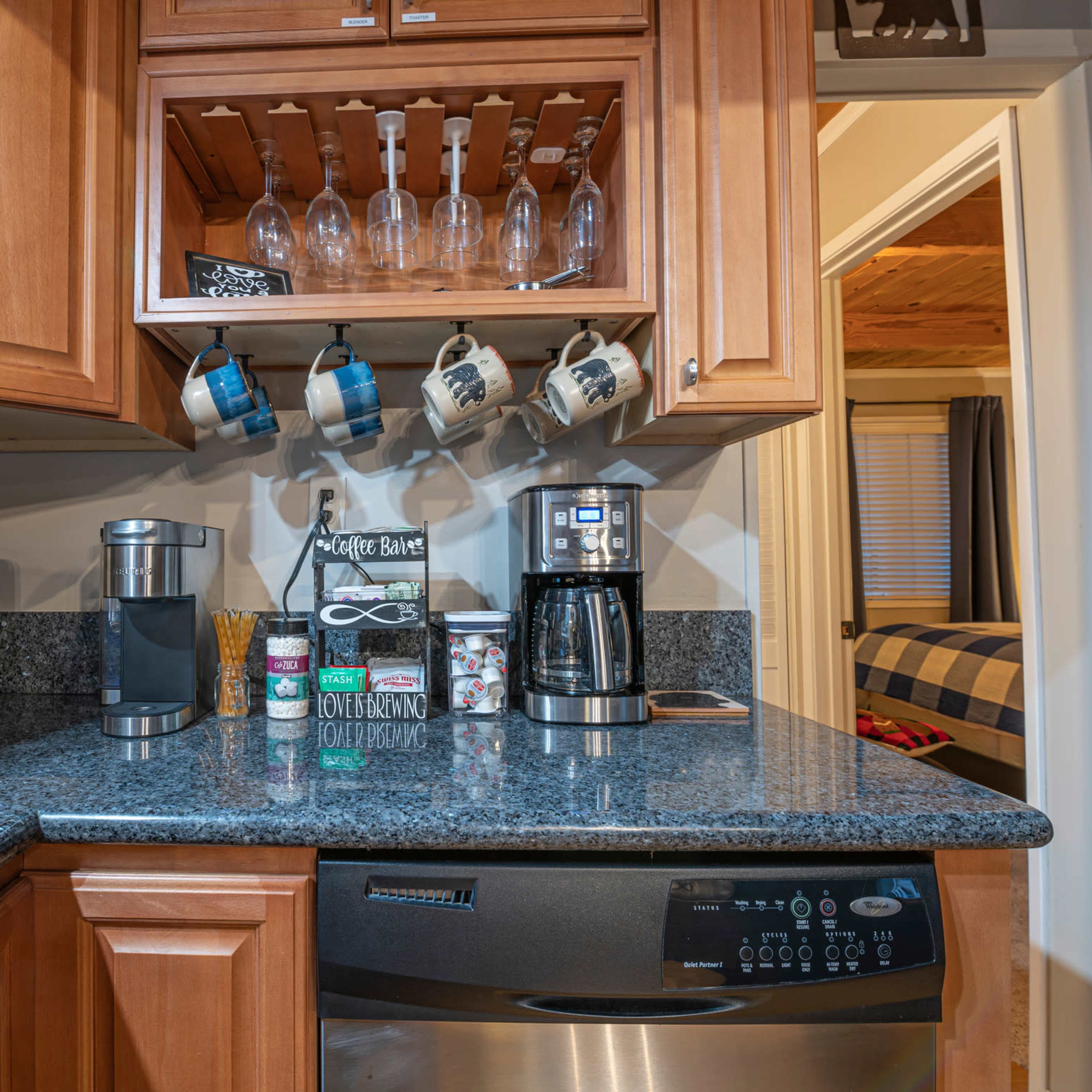 A kitchen countertop features a coffee maker, a blender, and hanging mugs, with glassware stored above and dark wood cabinetry in the background.