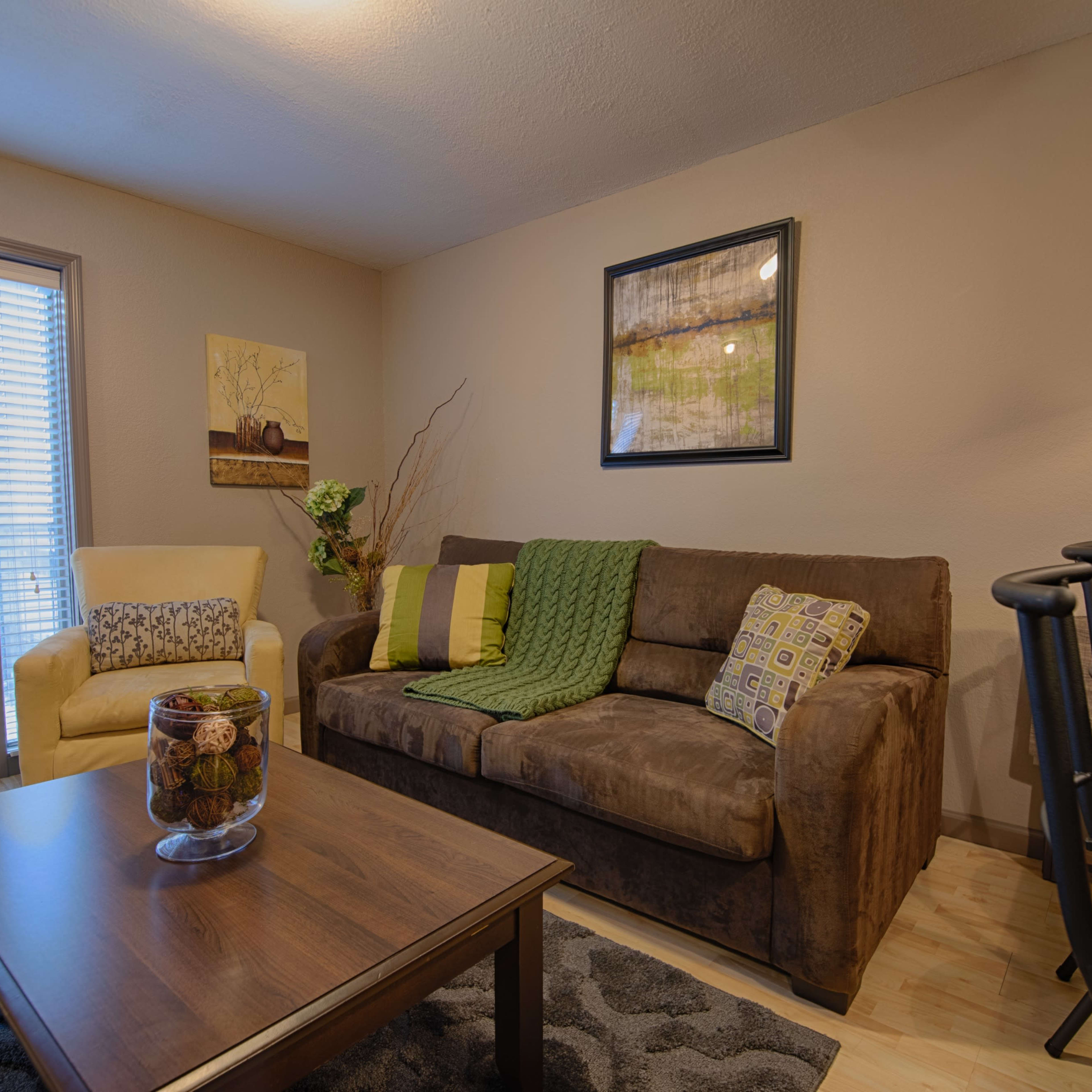 A living room with a brown couch adorned with green and patterned cushions, a wooden coffee table, and a small dining area featuring high stools.