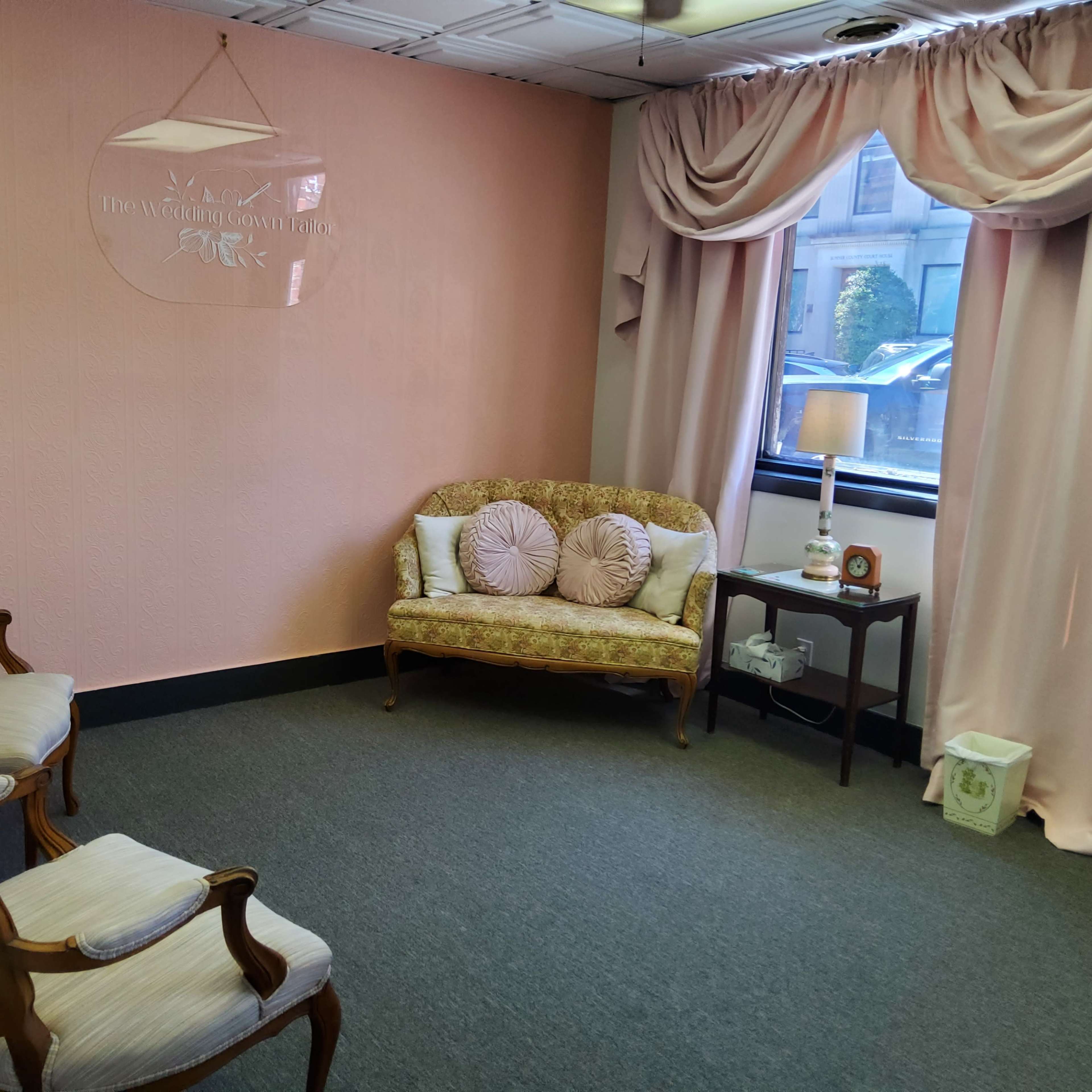 A waiting room with a pink wall, a patterned couch, two wooden chairs, a lamp, and a small table by a window.