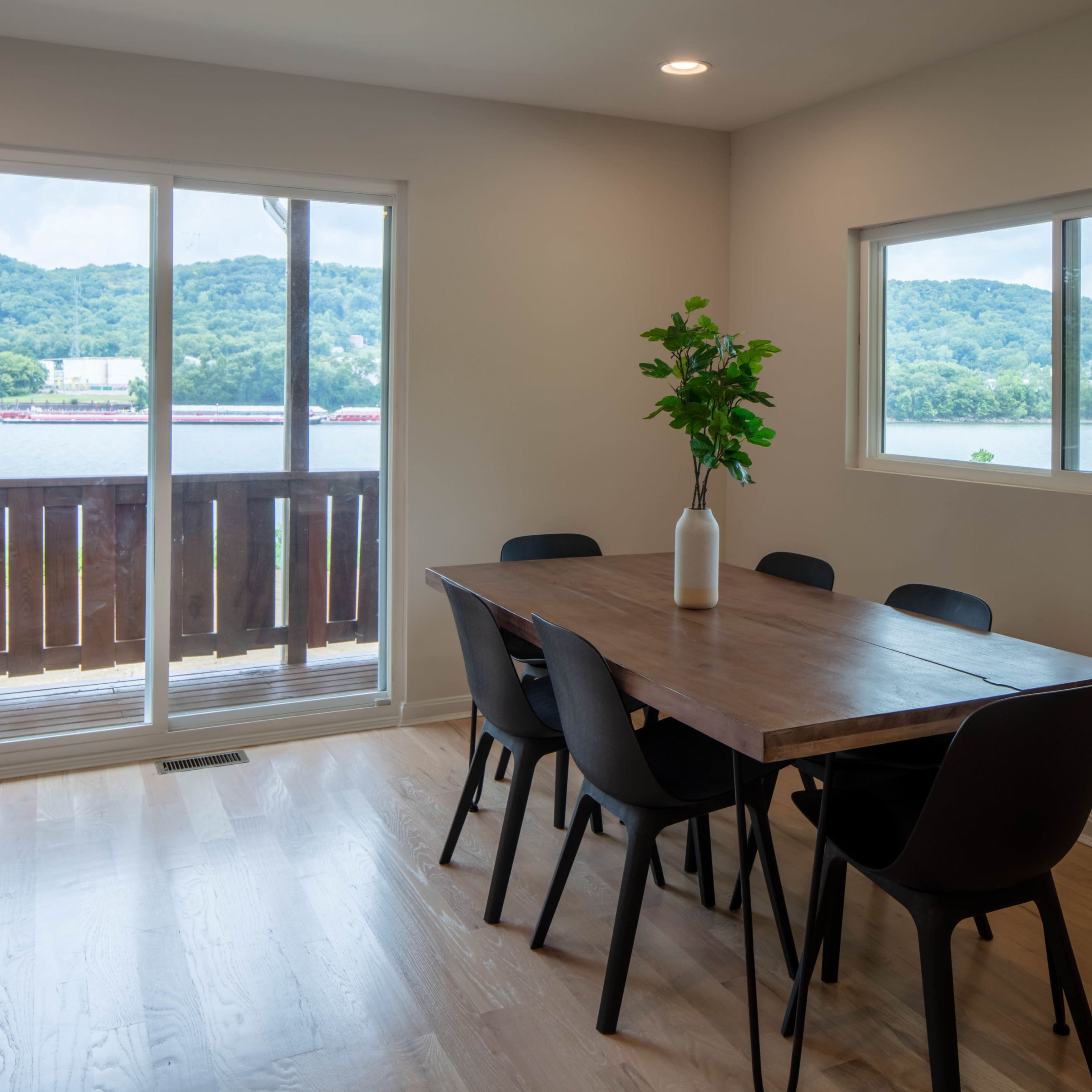 A dining area with a wooden table and black chairs overlooks a scenic view of hills and a river through large windows.