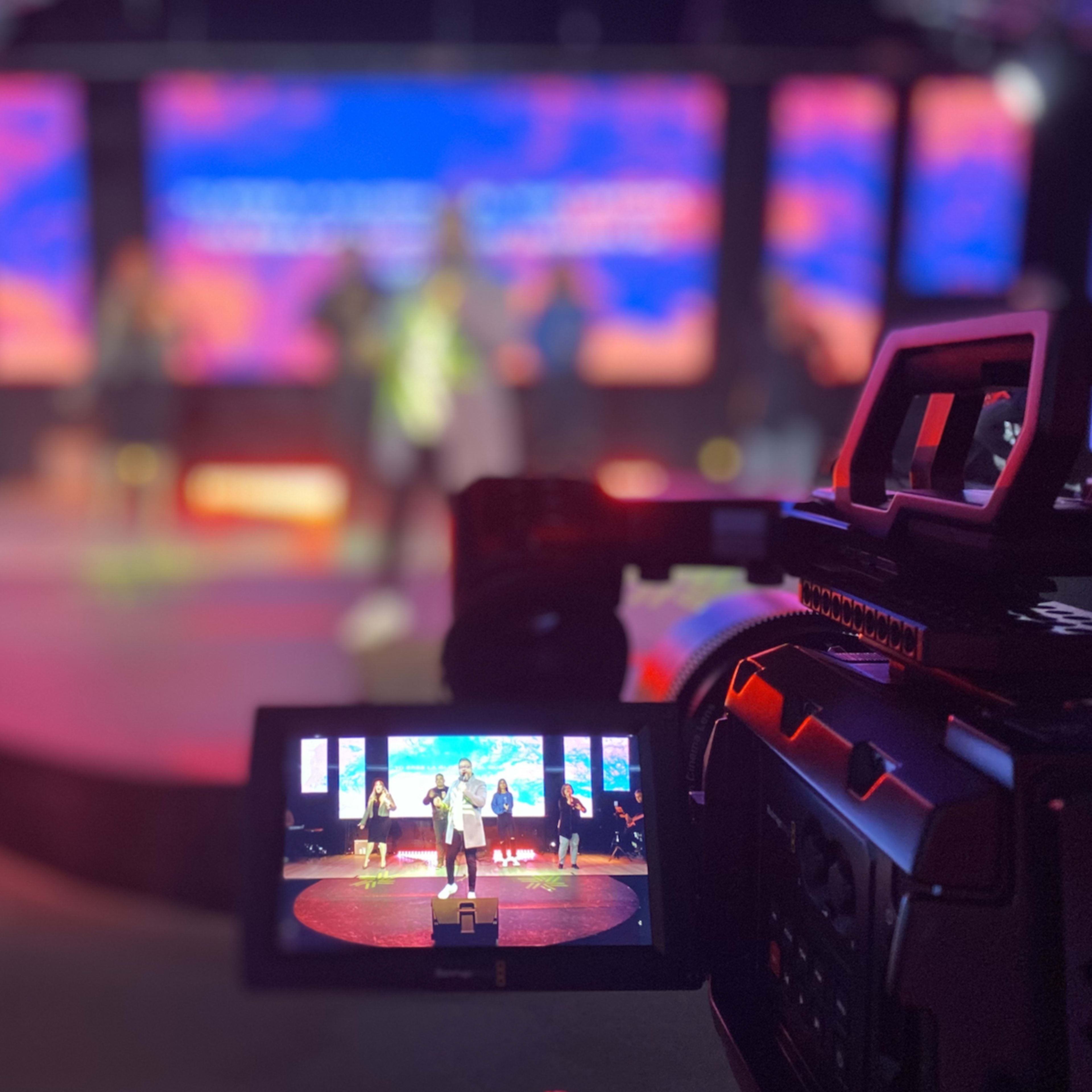 A video camera captures a performance on stage featuring multiple performers and a colorful backdrop.