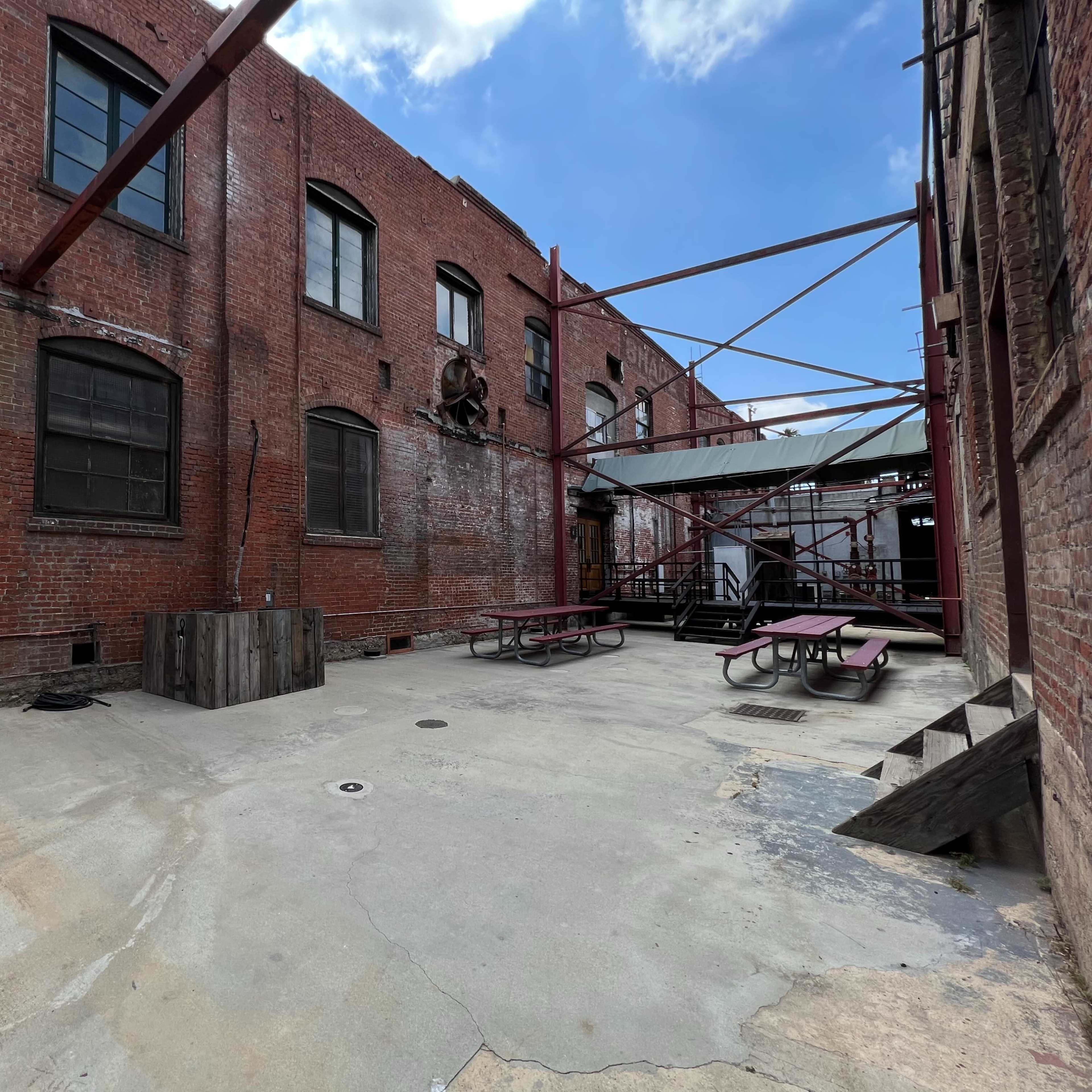 An empty, brick-walled courtyard with concrete flooring, picnic tables, and a metal support structure under a clear sky.
