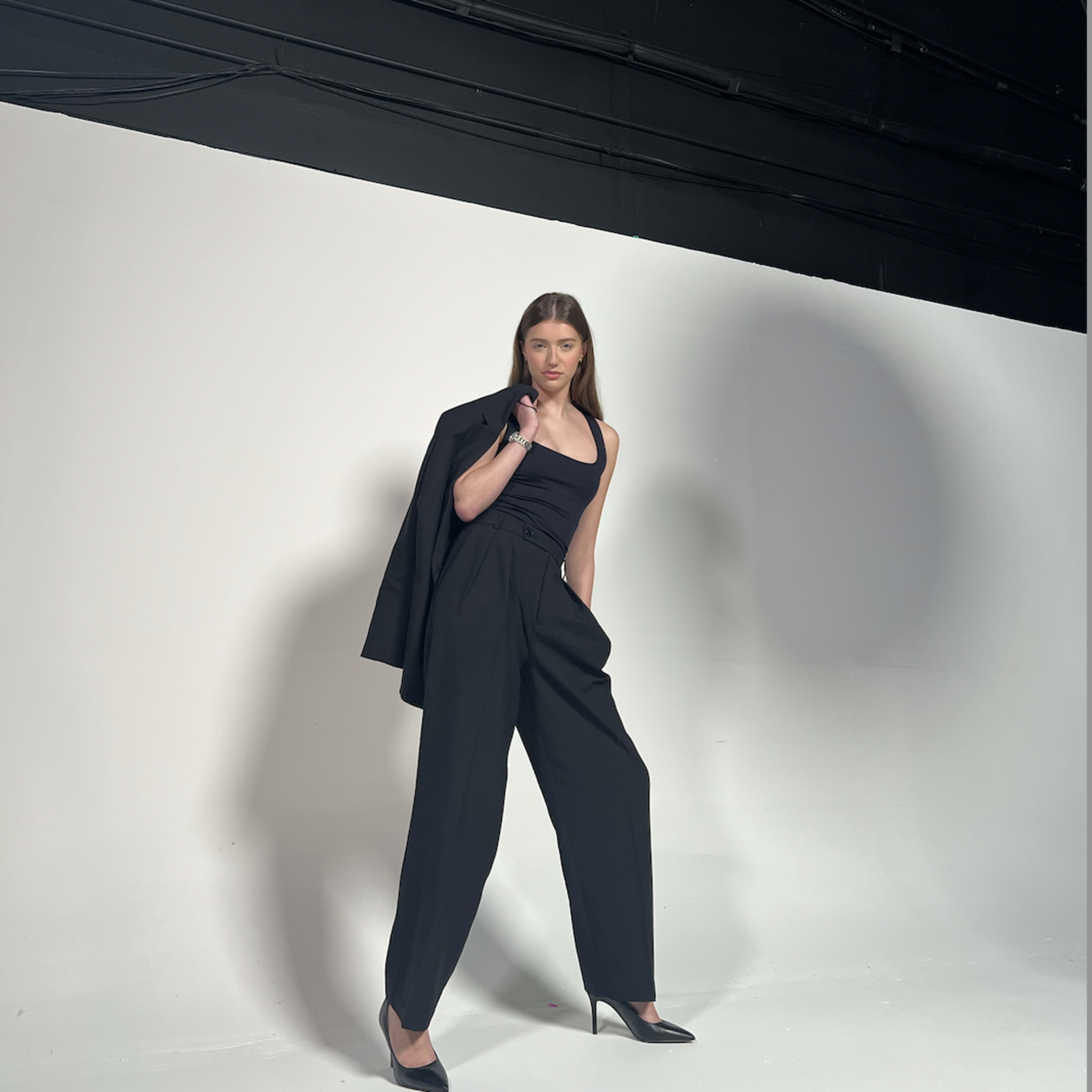 A model poses in a black outfit with a tailored blazer draped over one shoulder against a plain white background.