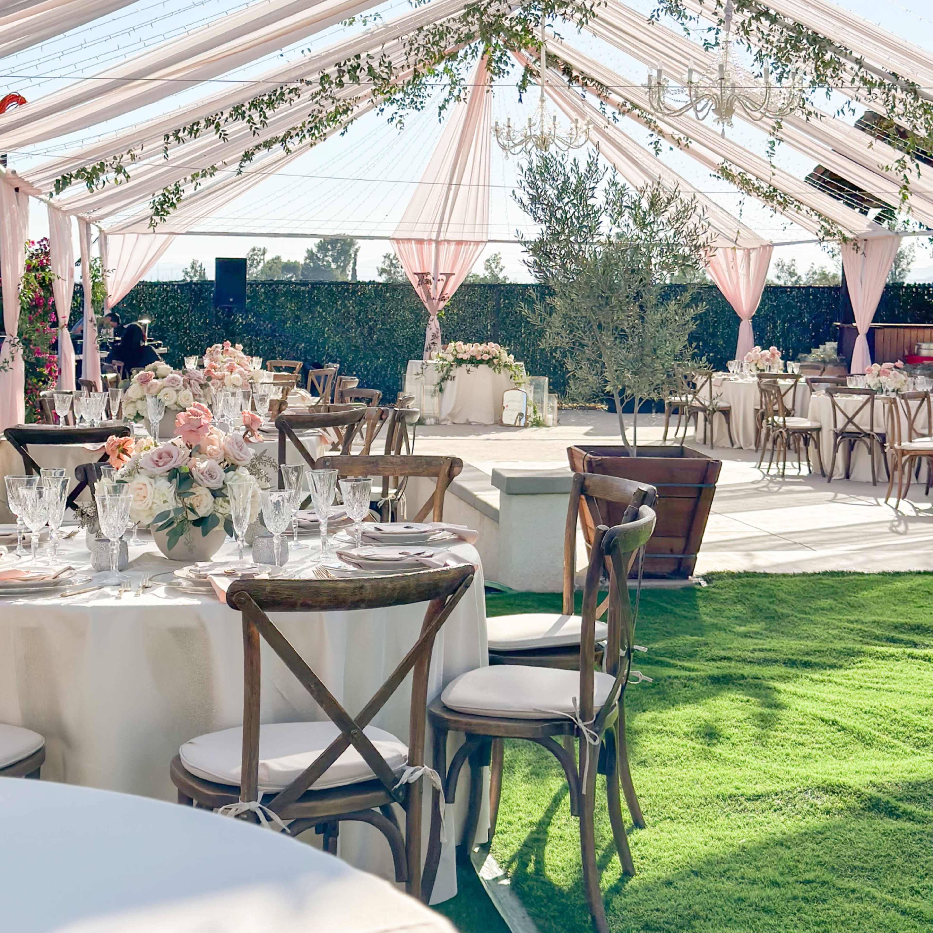 A tented outdoor reception area features elegantly set tables with floral centerpieces under draped fabric and string lights.