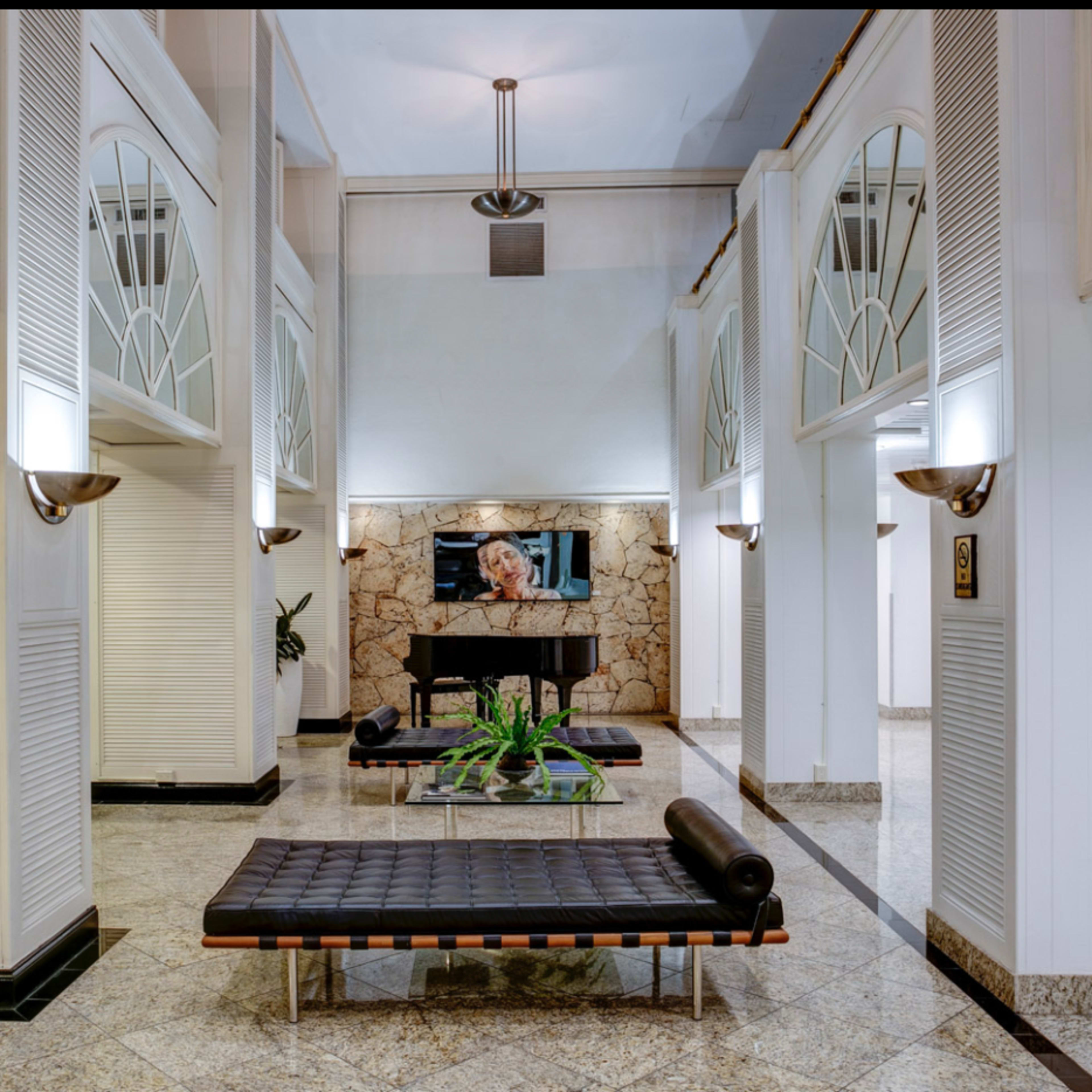 A spacious lobby area featuring a piano, benches, and decorative plants, with large windows allowing natural light to enter.