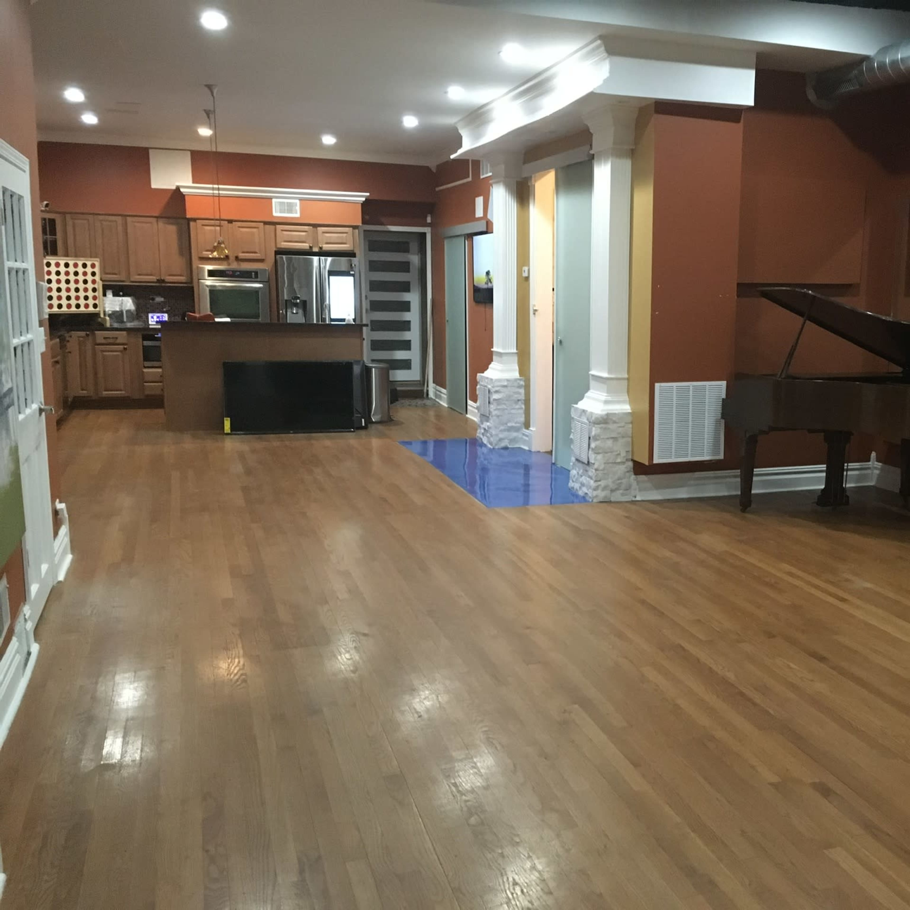 A spacious interior room with hardwood floors, featuring a kitchen area on the left, a grand piano near the center, and columns along the walls.