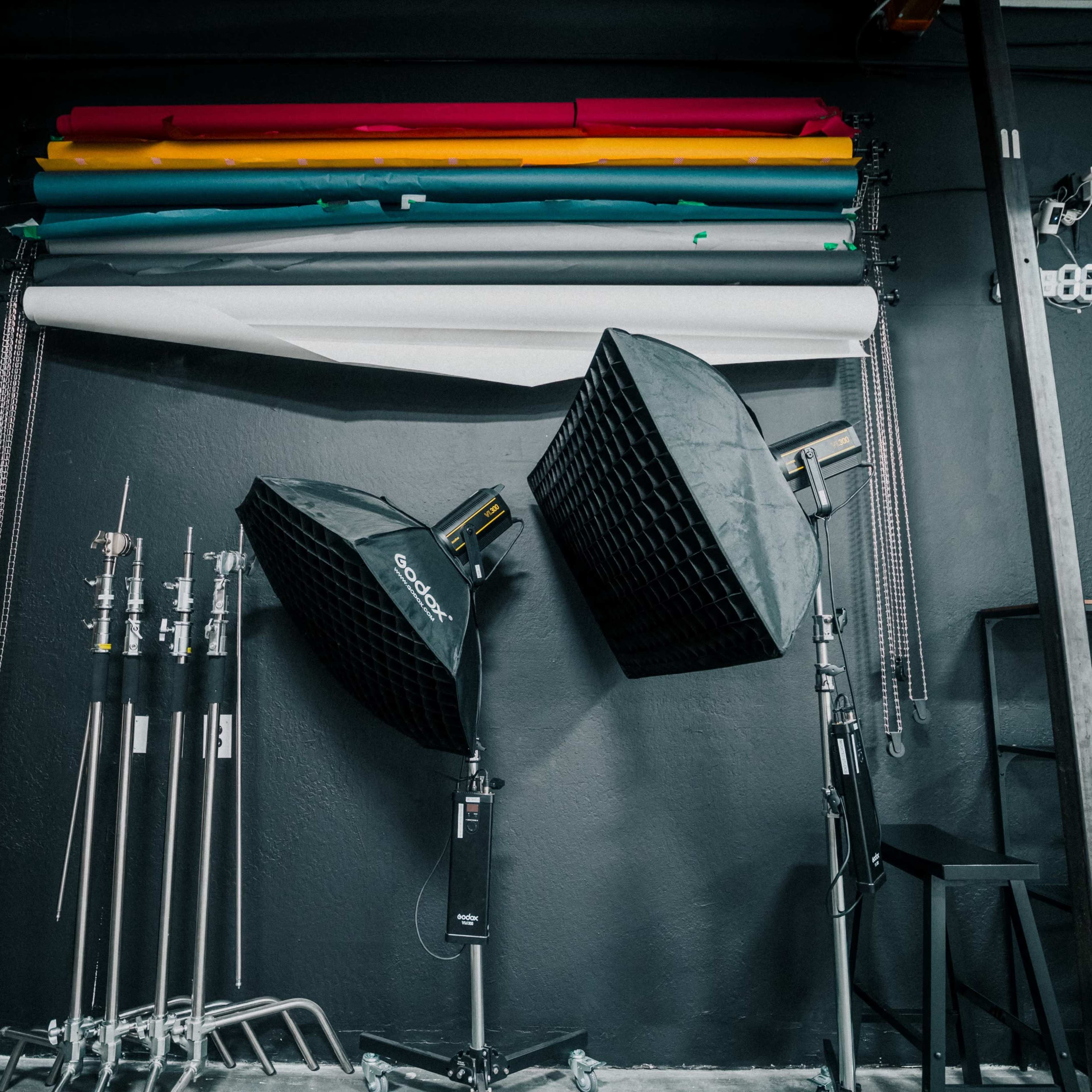 A photography studio with backdrop rolls in various colors hanging on the wall and two softbox lights positioned on stands.