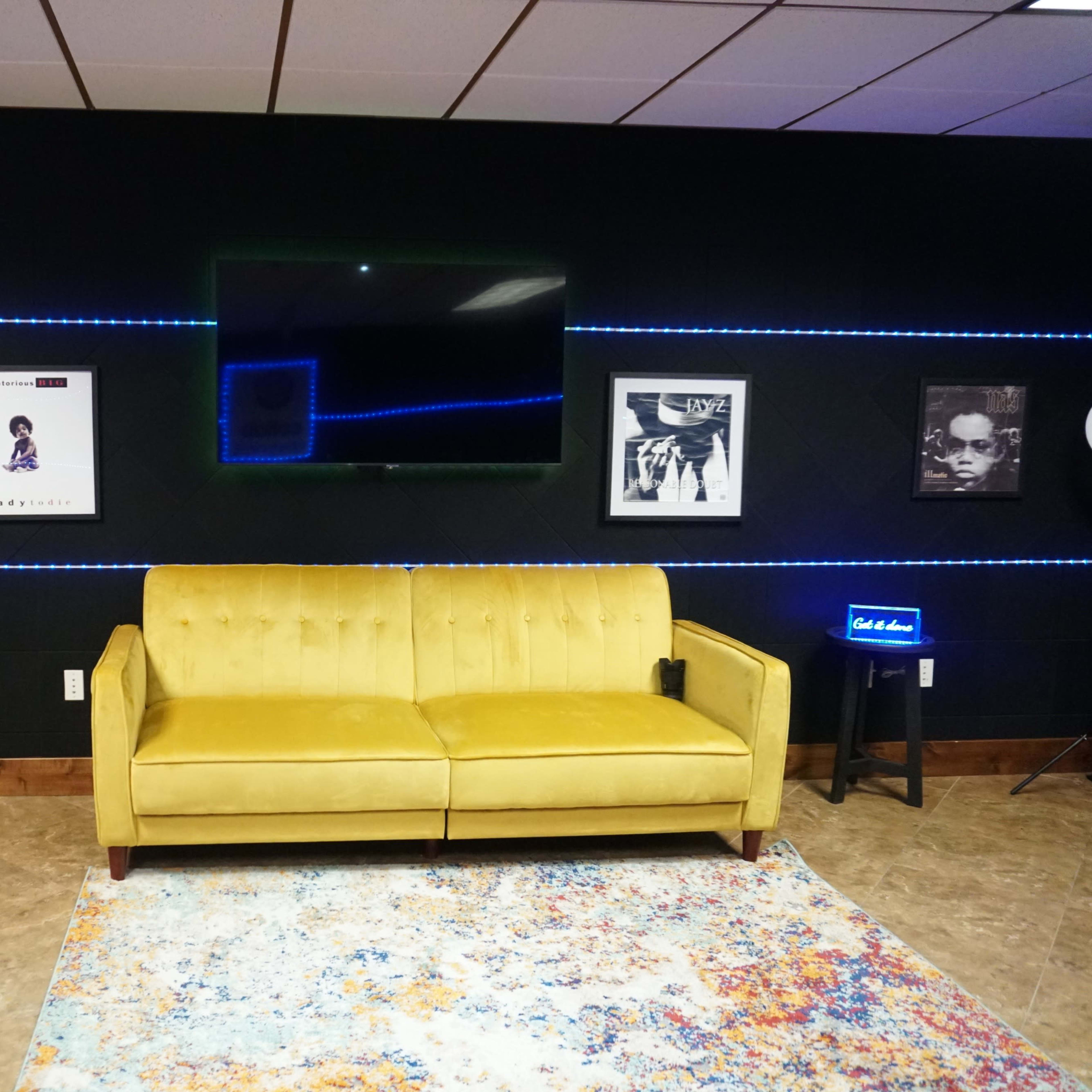 A yellow sofa positioned against a black wall adorned with framed posters and illuminated by blue LED strip lights.