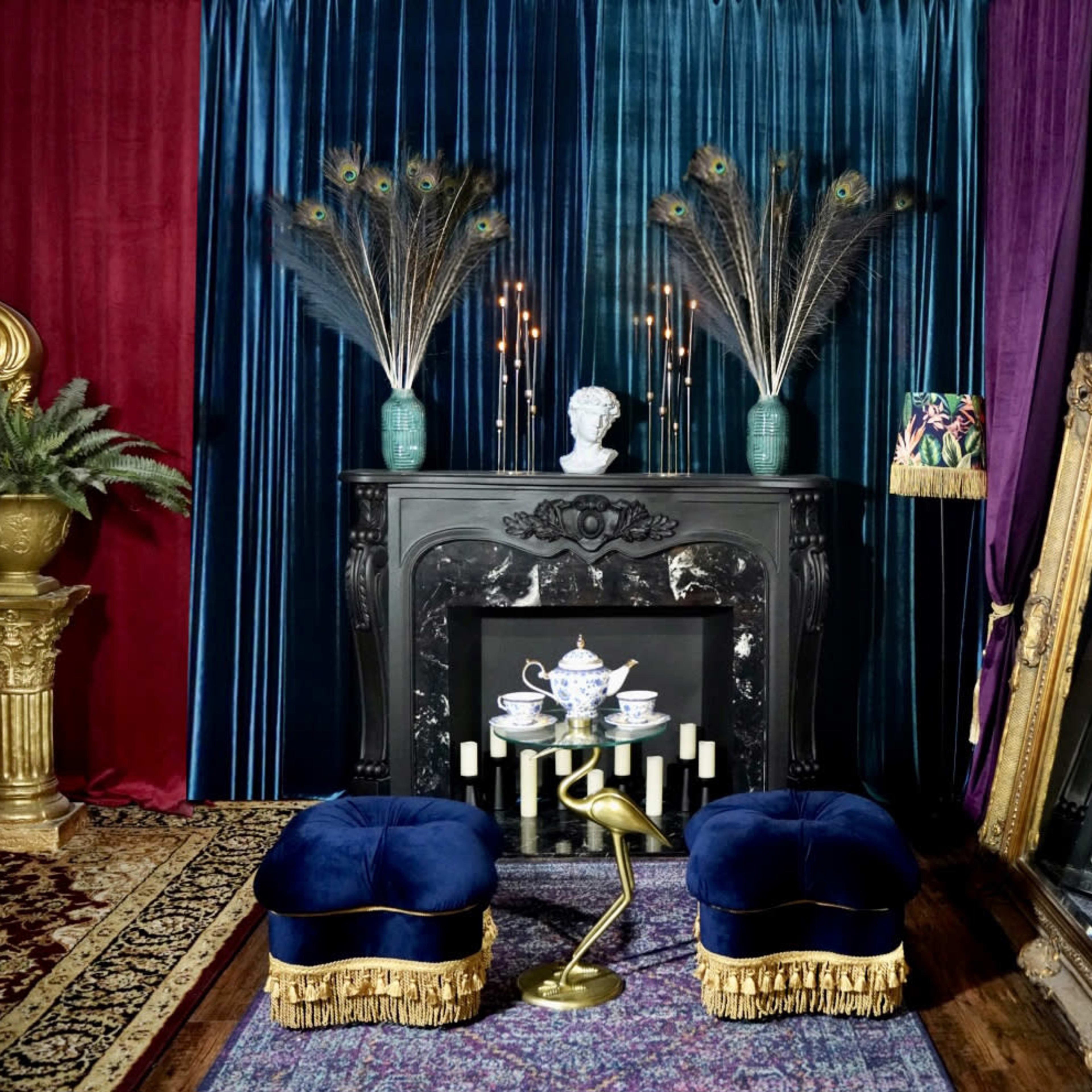 The scene features a richly decorated interior with a dark fireplace adorned by a bust, peacock feathers in vases, and two blue velvet stools in front of an ornate mirror.