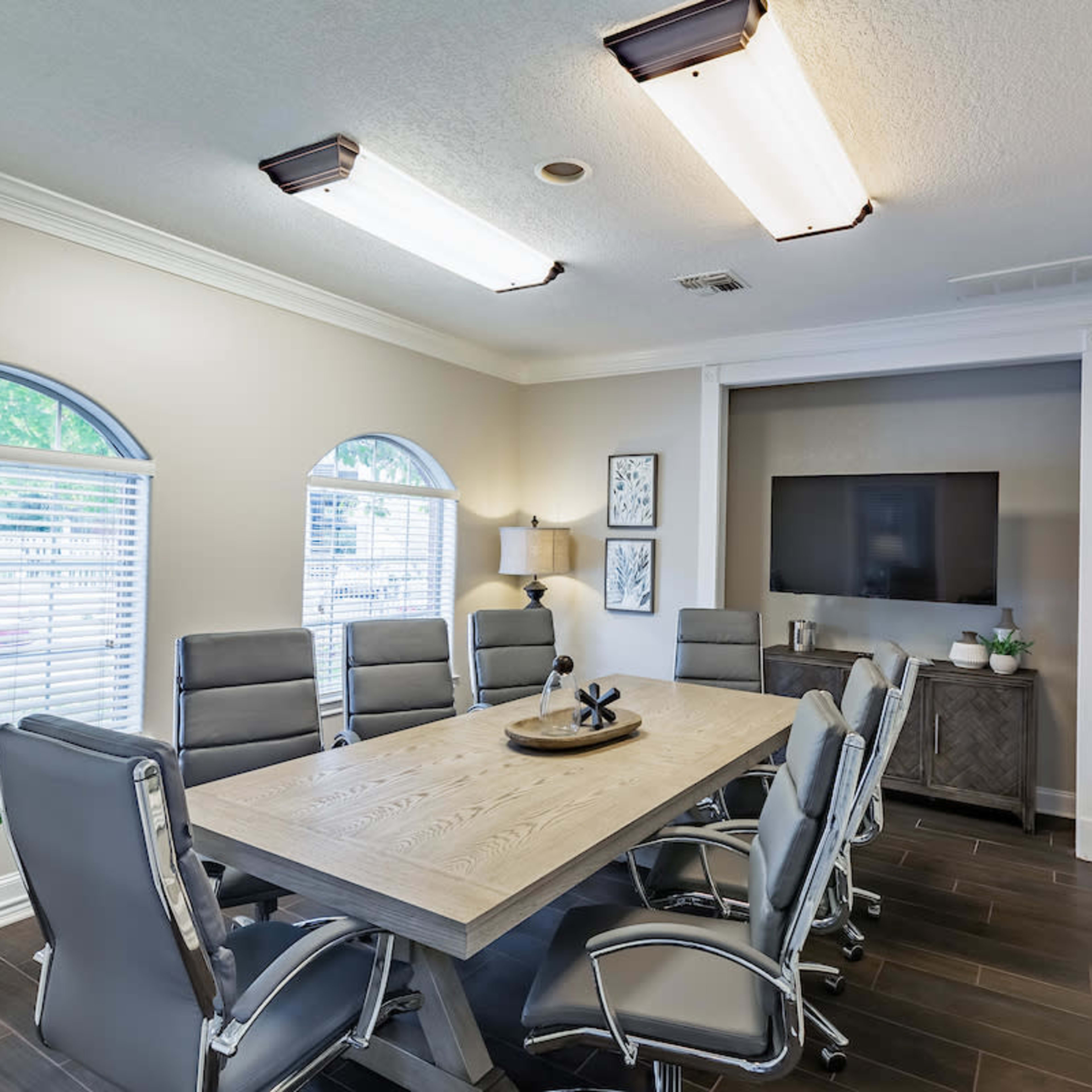 A conference room features a large wooden table surrounded by six gray chairs, with a television on the wall and natural light coming through arched windows.