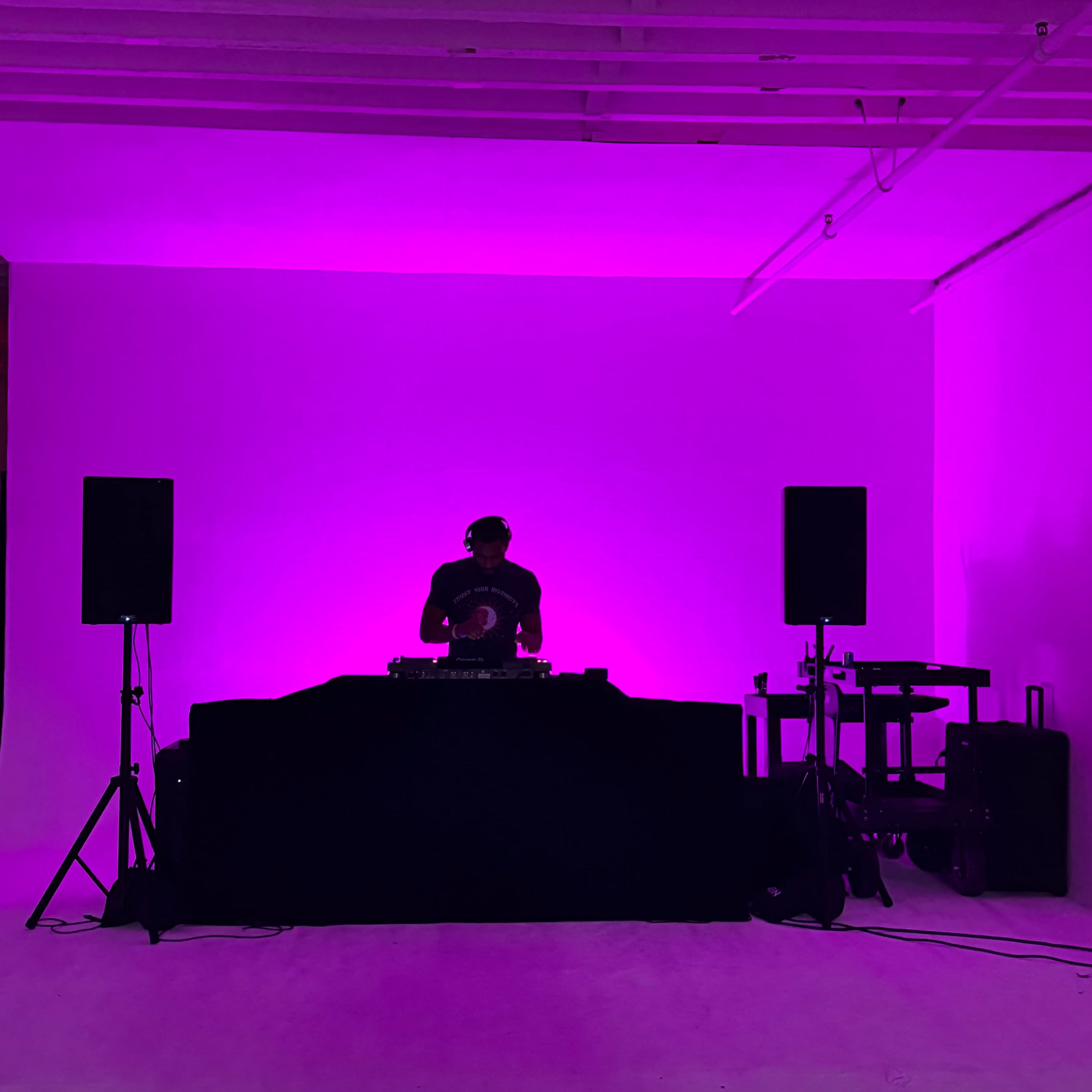 A DJ stands behind a mixer in a room illuminated with pink light, flanked by two speakers.