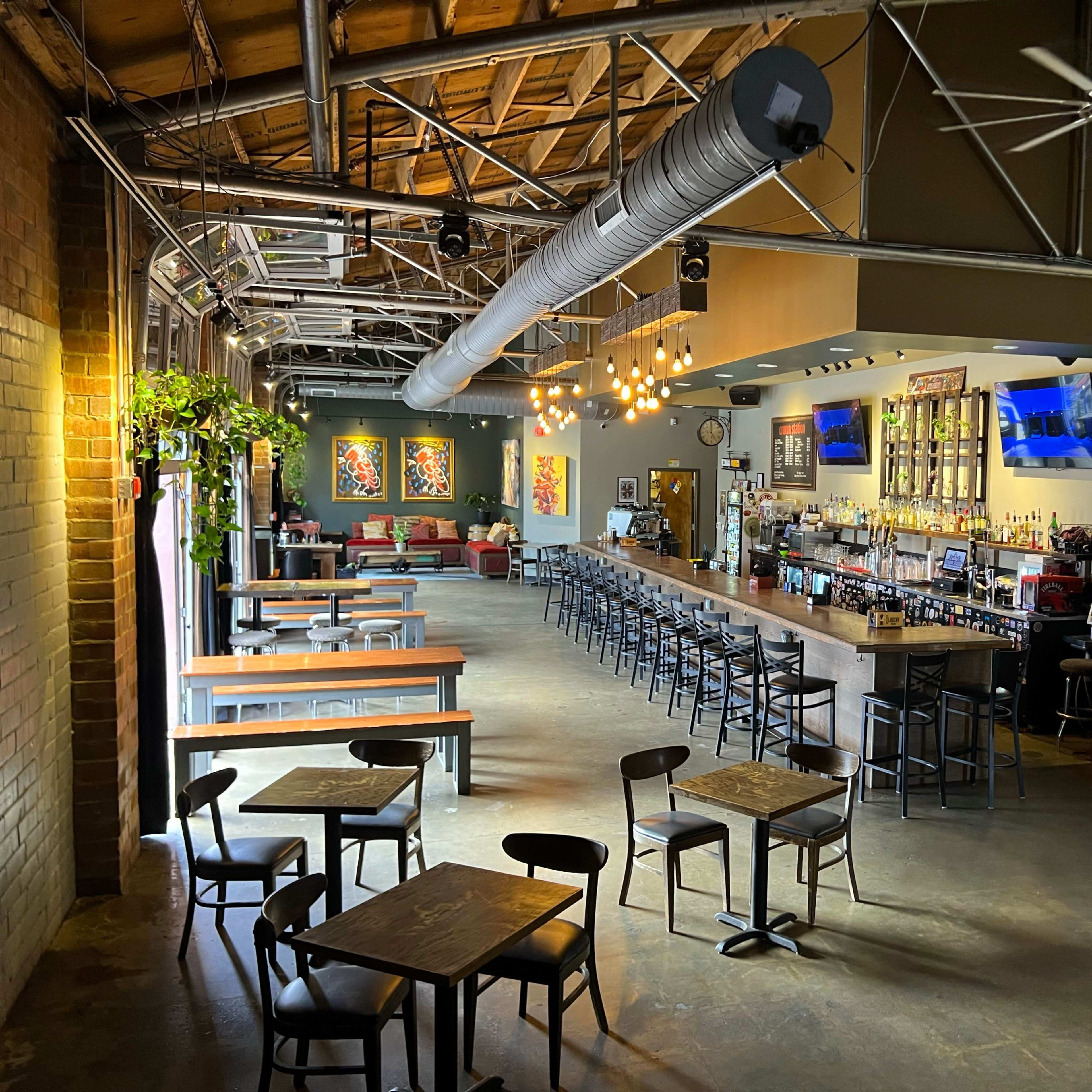 A spacious bar and dining area with a long wooden counter, rows of black stools, and tables scattered throughout, illuminated by pendant lights and adorned with colorful artwork on the walls.