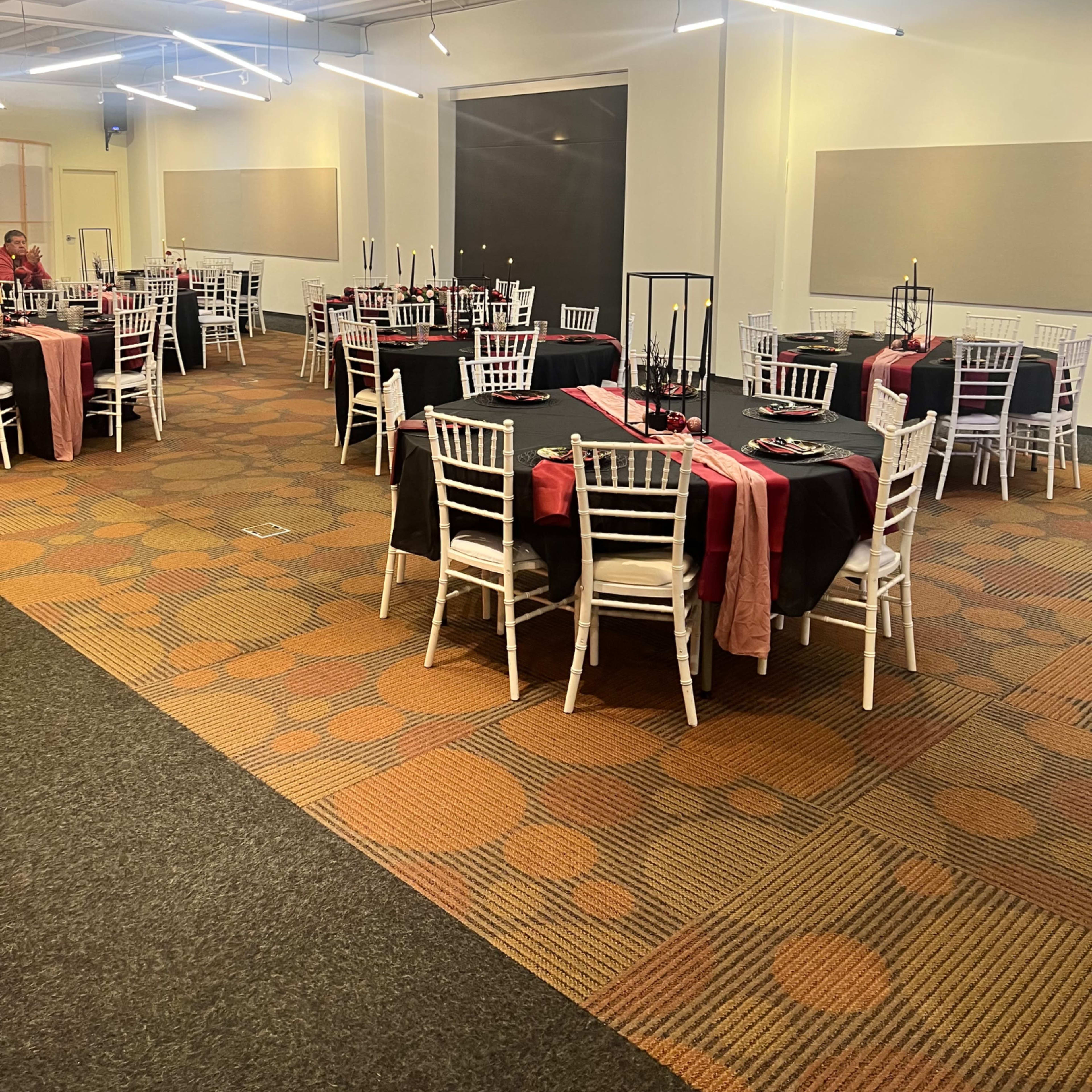A spacious event room set with several round tables covered in black and red tablecloths, surrounded by white chairs.