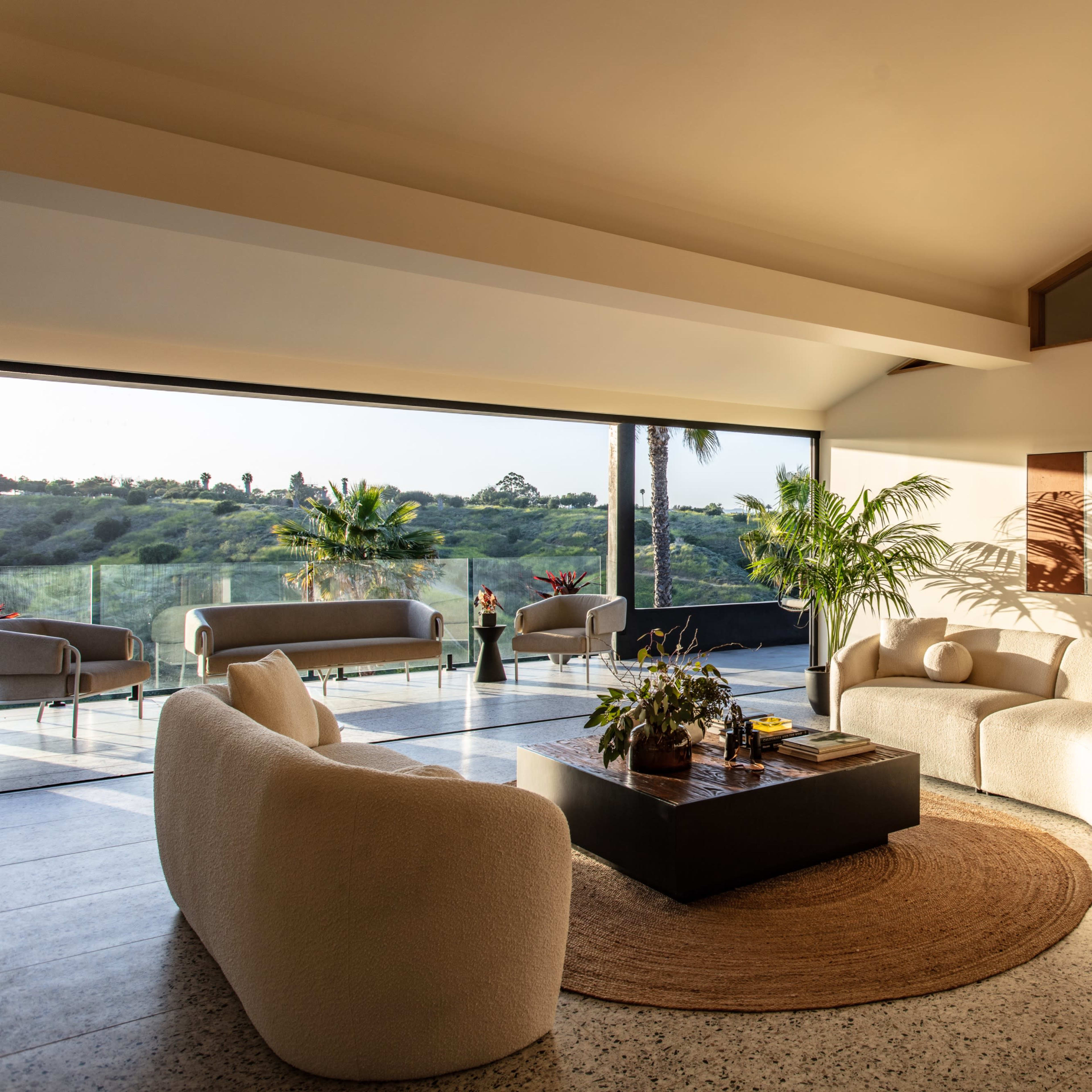 A modern living room features a curved sectional sofa, a round area rug, and a coffee table, with large windows offering a view of greenery outside.