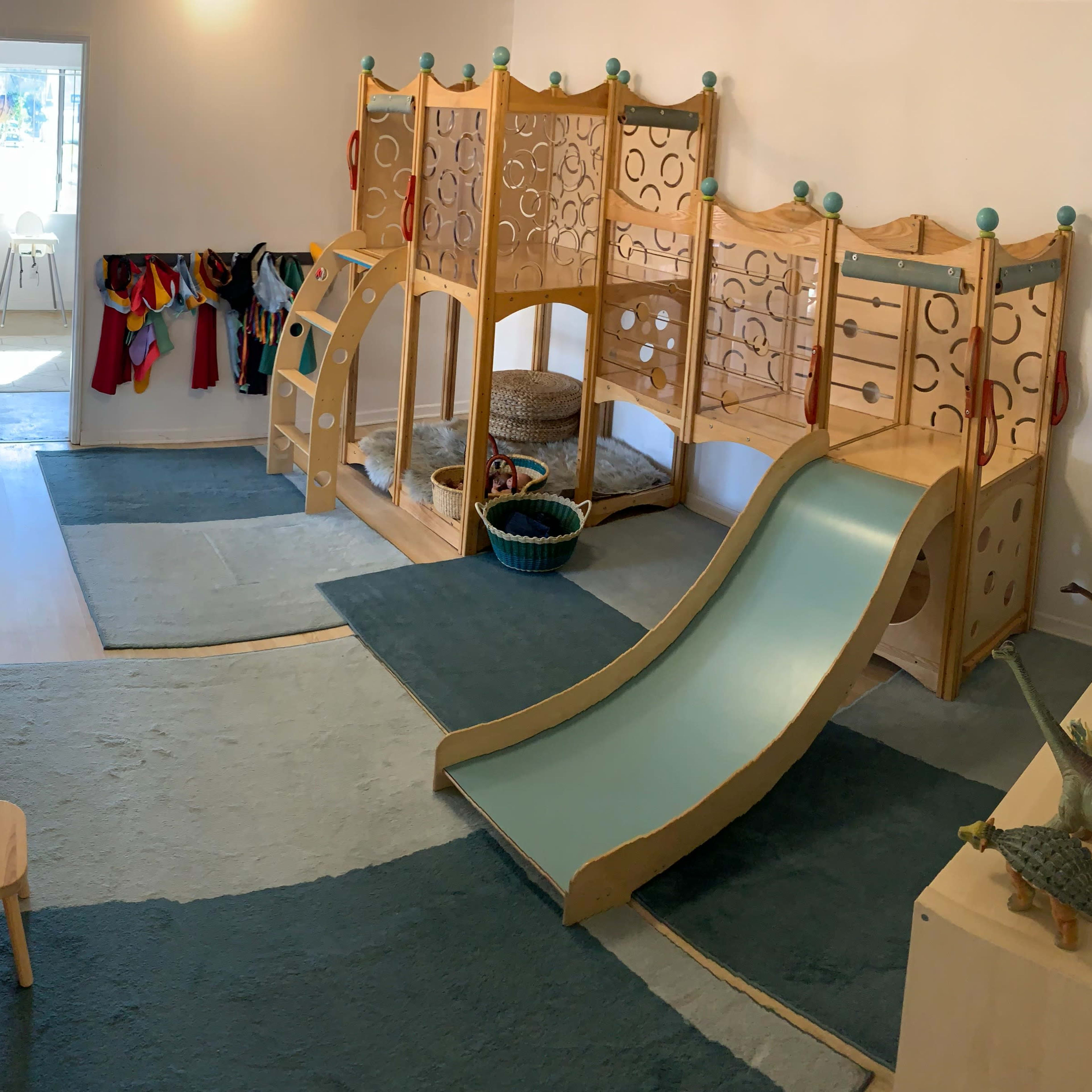 A brightly lit playroom featuring a wooden climber with a slide, soft blue rugs, and a small table with plates in one corner.