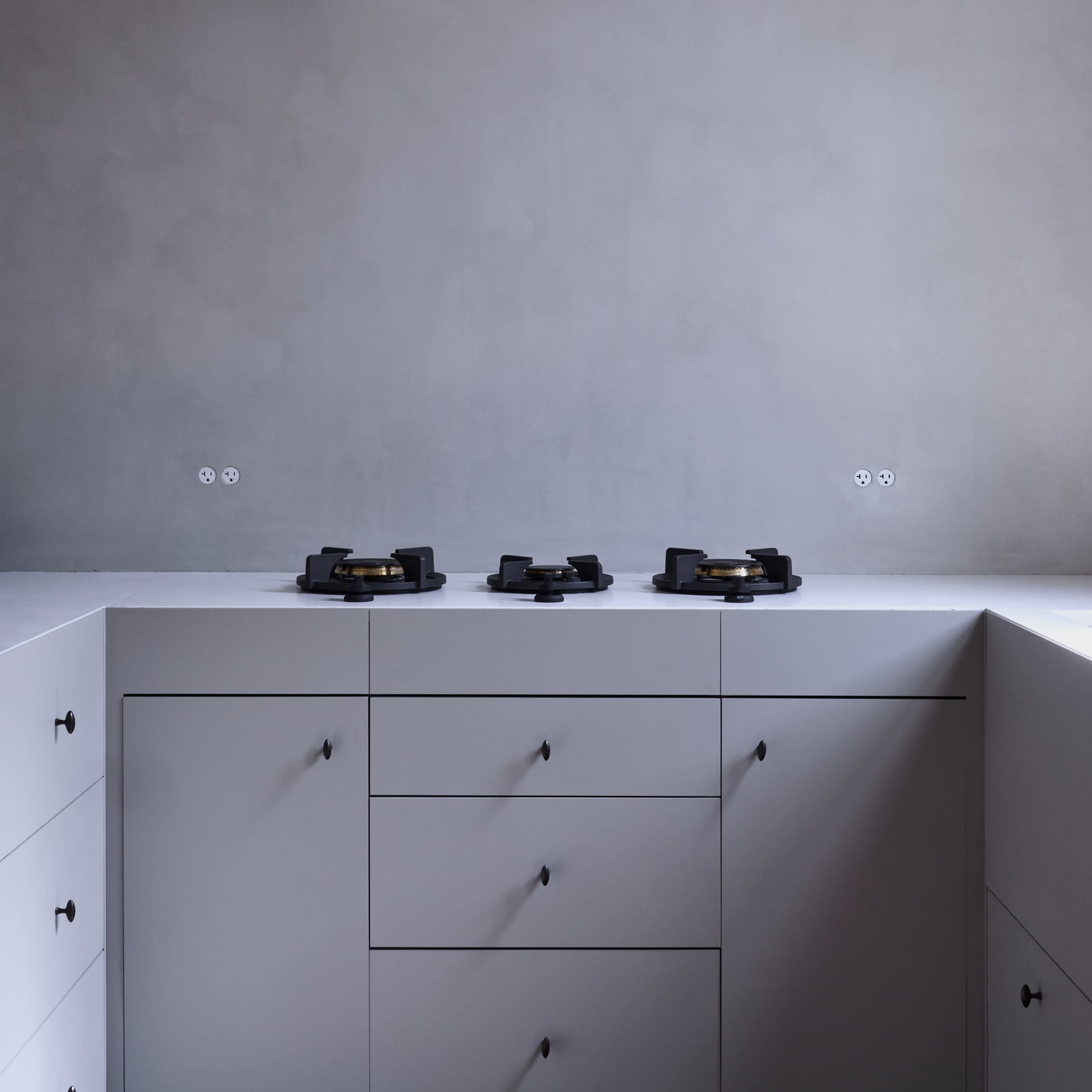 A modern kitchen features a minimalist design with gray walls and a countertop equipped with three black cooking burners.