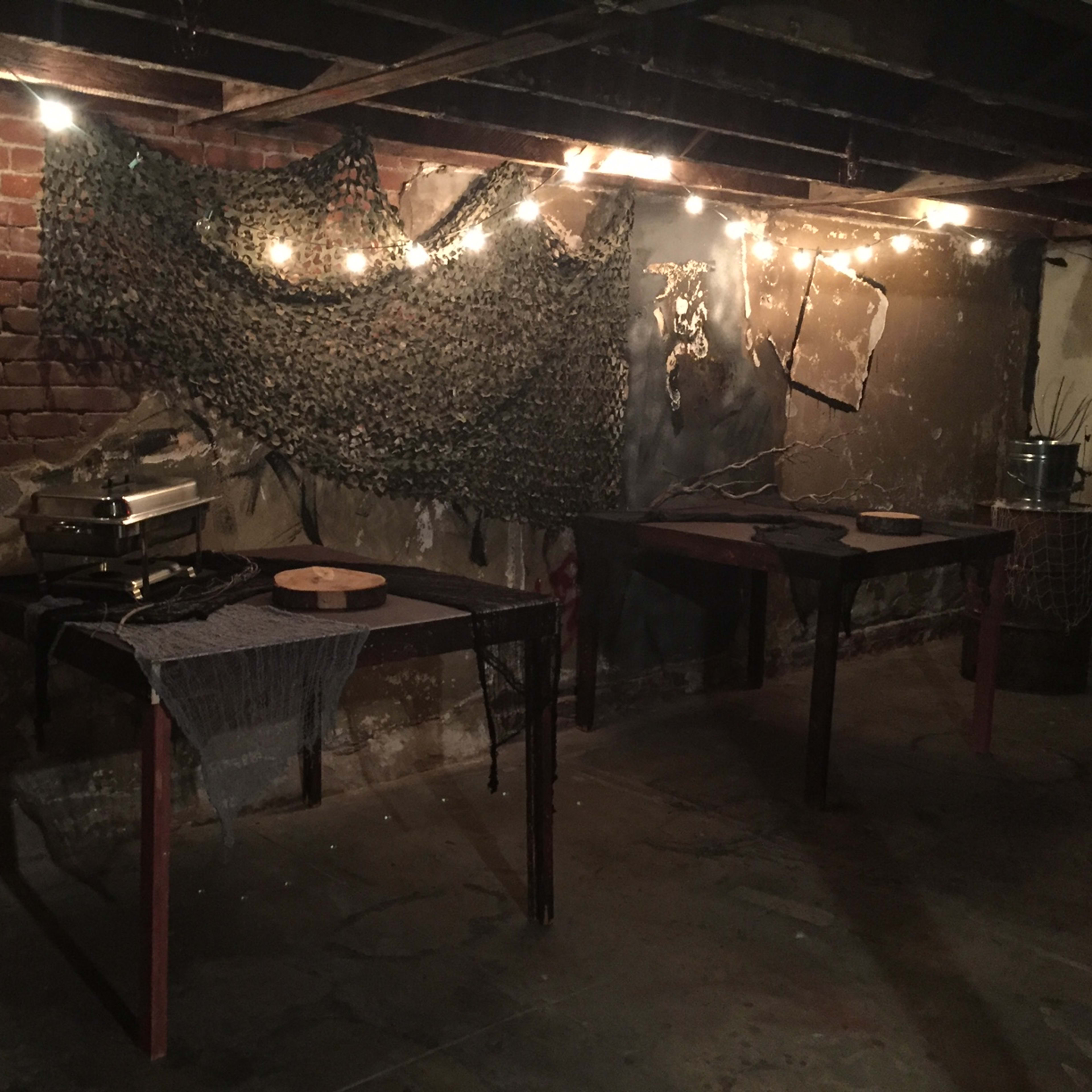 A dimly lit basement with two wooden tables, a backdrop of camouflage netting, and string lights overhead illuminating the brick and concrete walls.