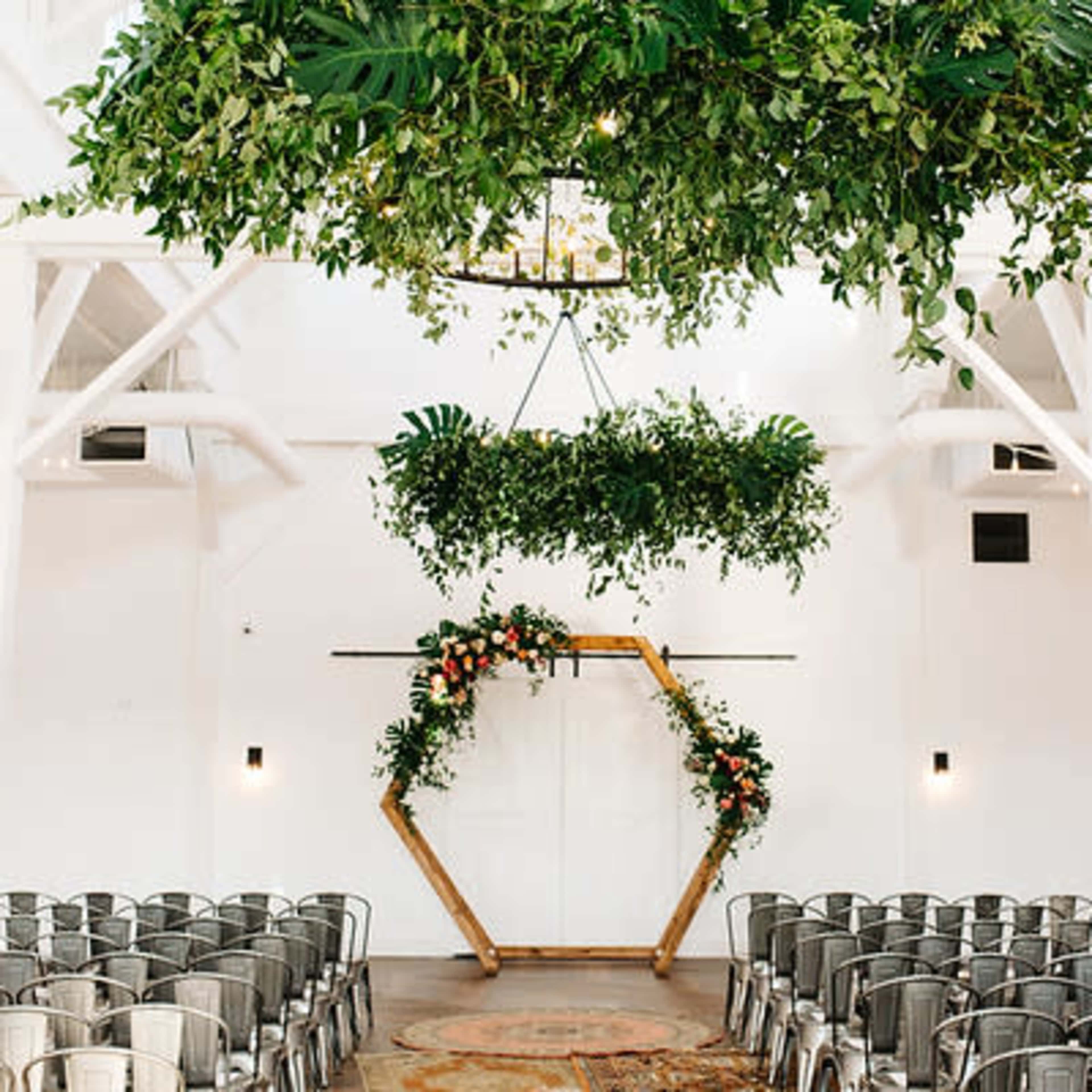 An indoor wedding venue features a hexagonal altar adorned with greenery and flowers, surrounded by rows of metal chairs under hanging plants.
