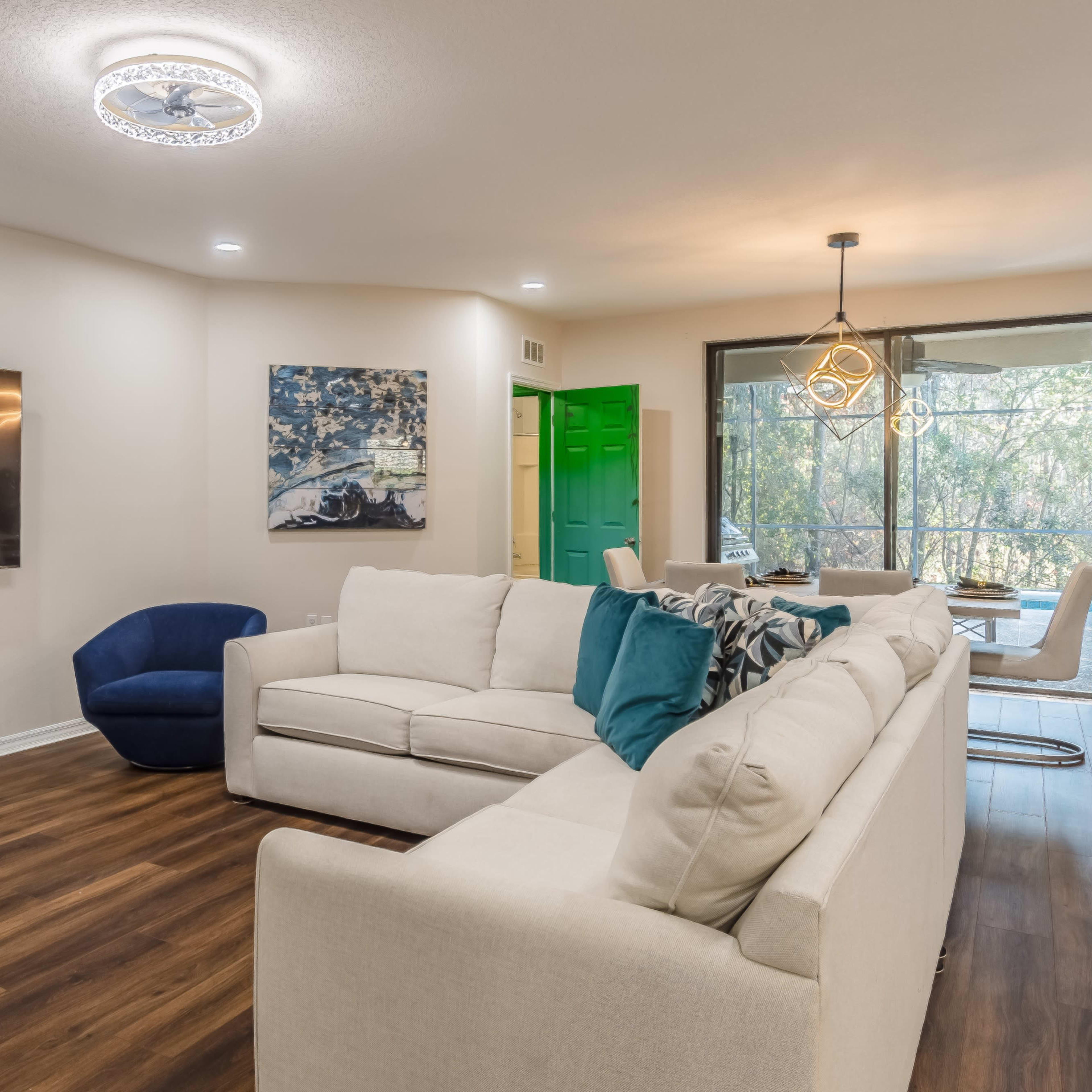 A modern living room features a light-colored sectional sofa, a dark blue accent chair, and a wall-mounted television, with large windows providing views of greenery outside.