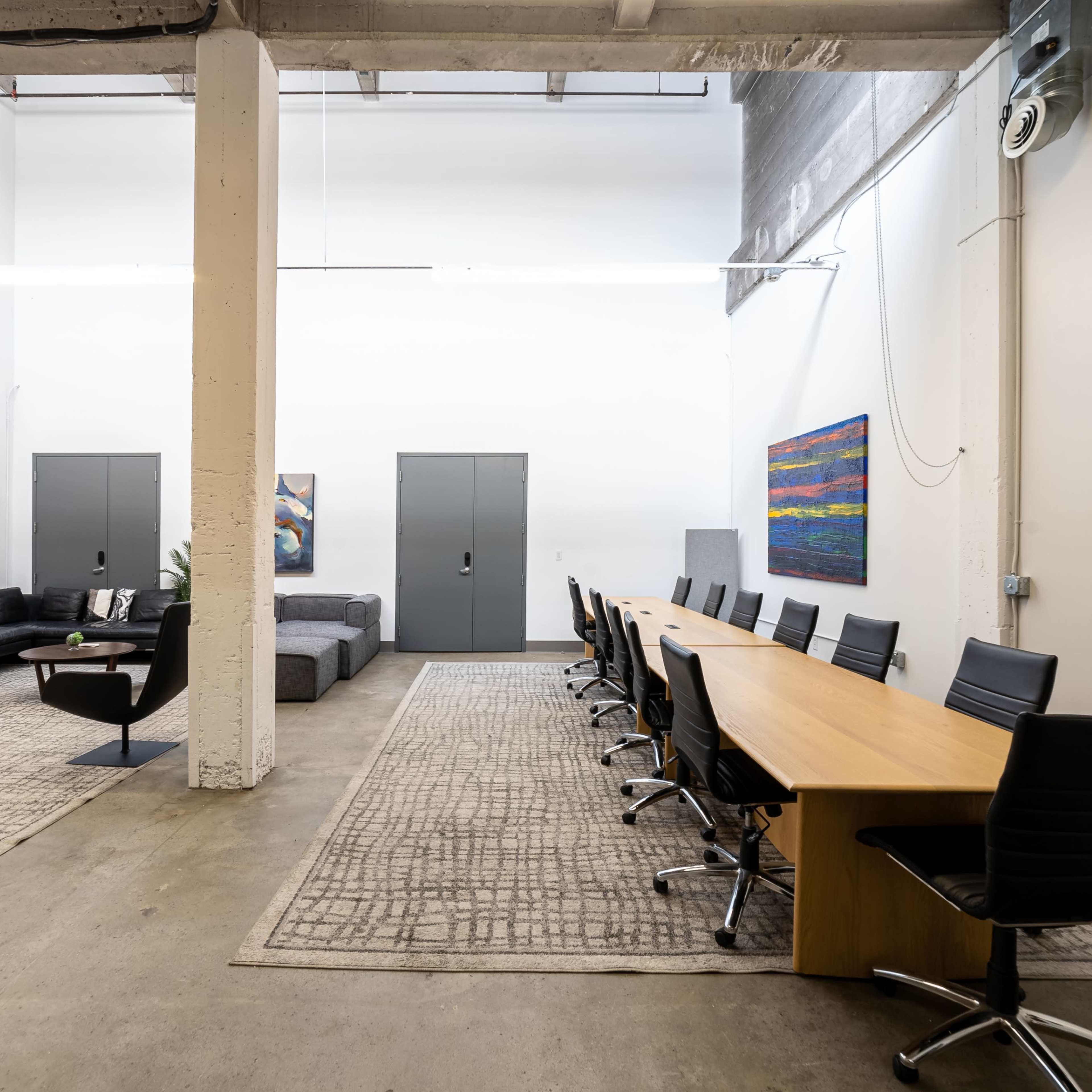 A spacious office interior featuring a long wooden conference table surrounded by black chairs, with a lounge area containing a dark couch and colorful artworks on the walls.