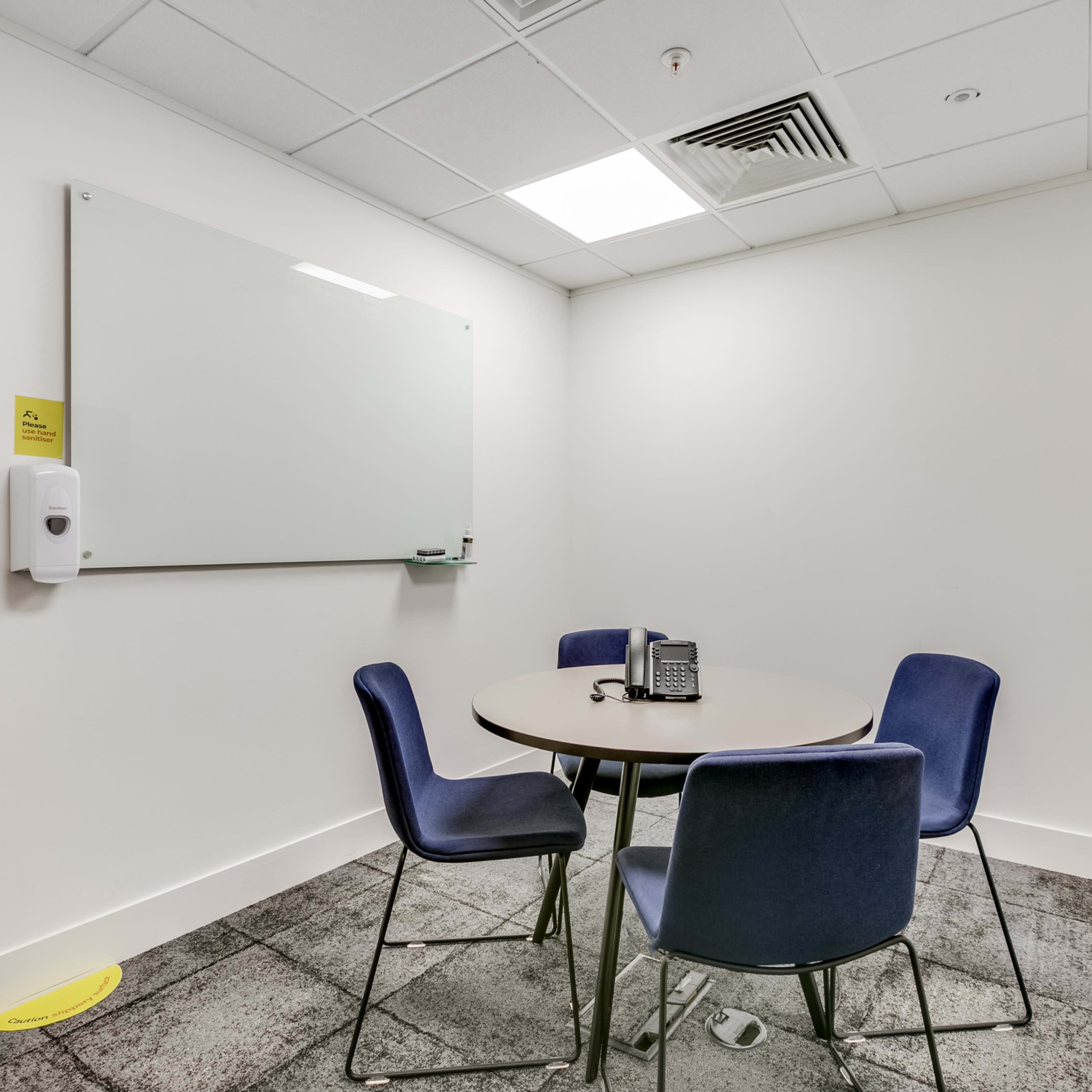 A small meeting room featuring a round table with four blue chairs, a whiteboard, a wall-mounted TV, a phone, and a hand sanitizer dispenser.