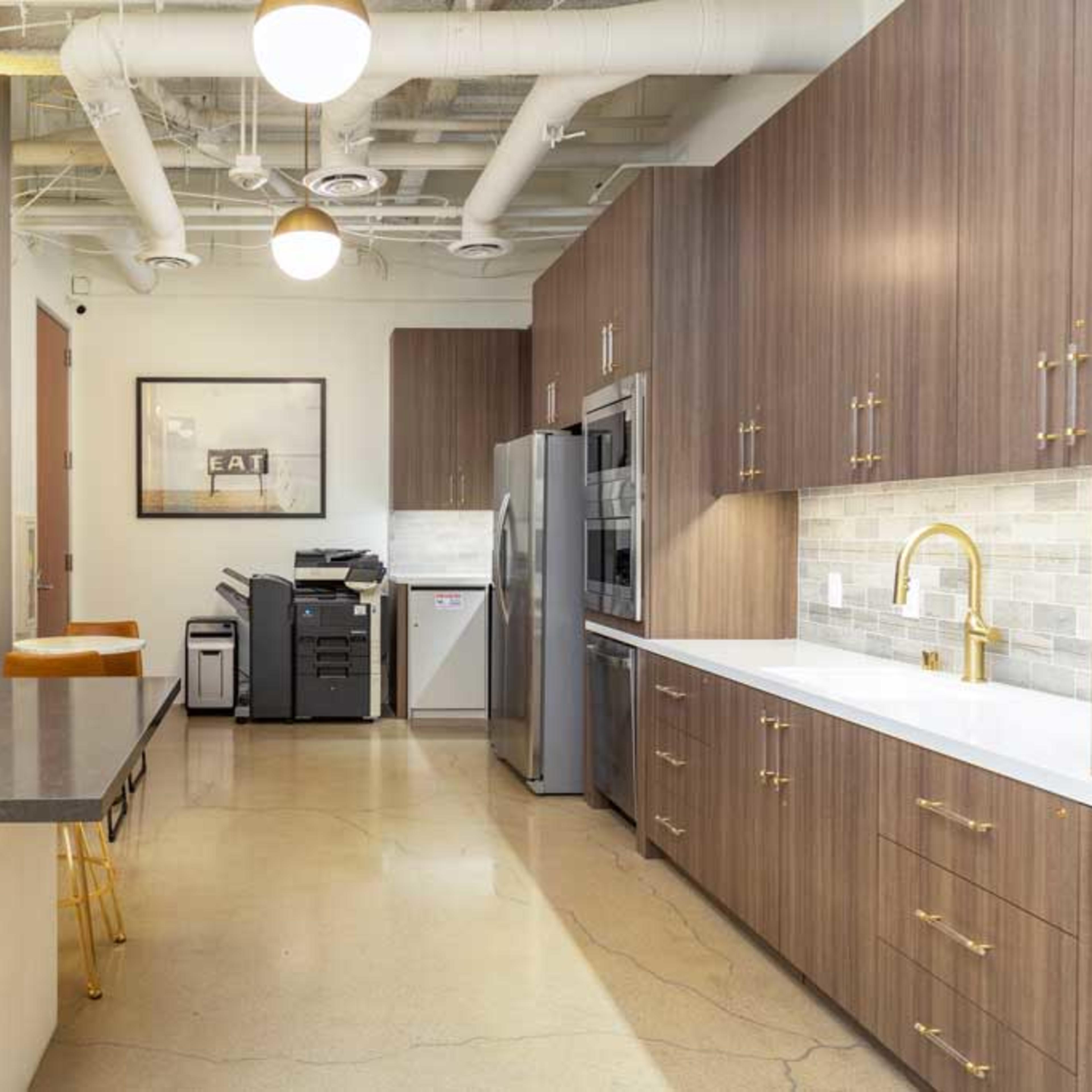 A modern kitchen with wooden cabinets, a large island with bar stools, and a small office space featuring a printer and a framed wall art.