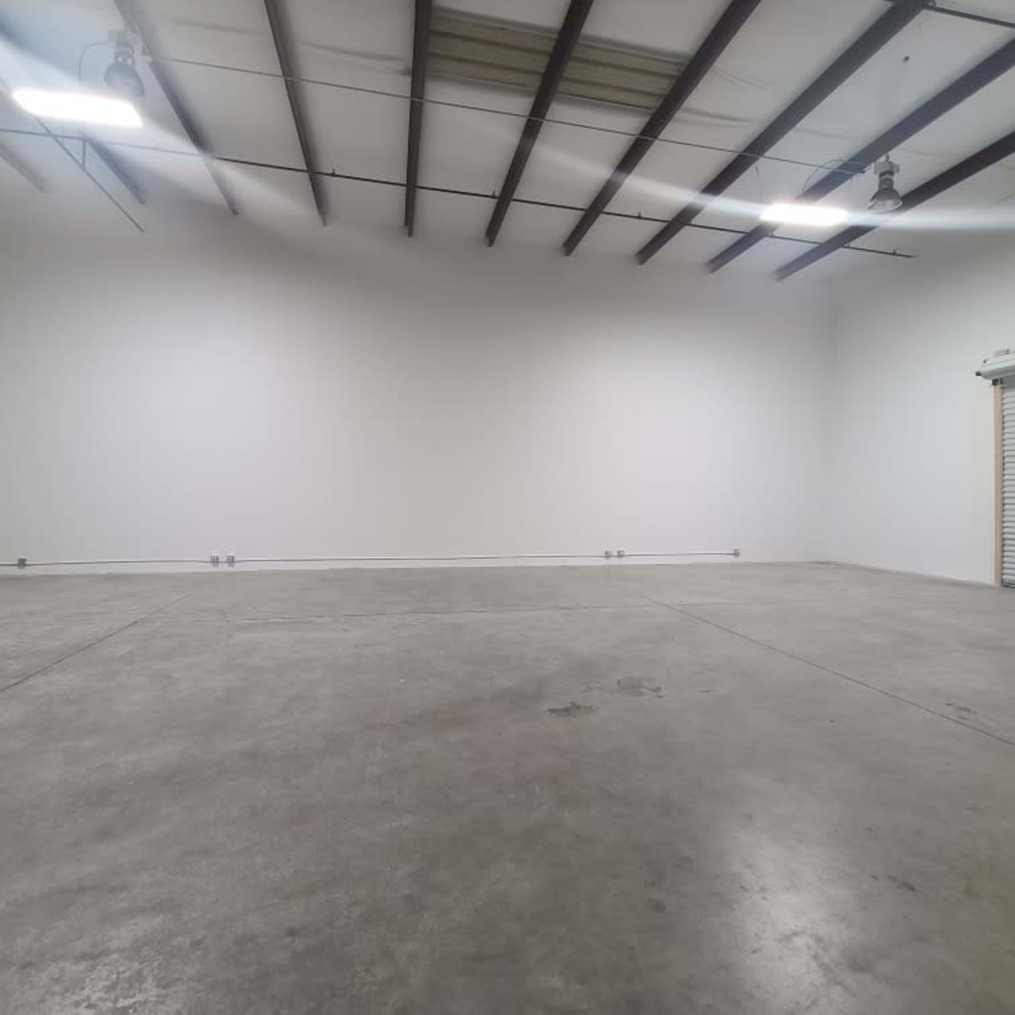 A spacious, empty industrial interior with concrete flooring, high ceilings, and a roll-up garage door.