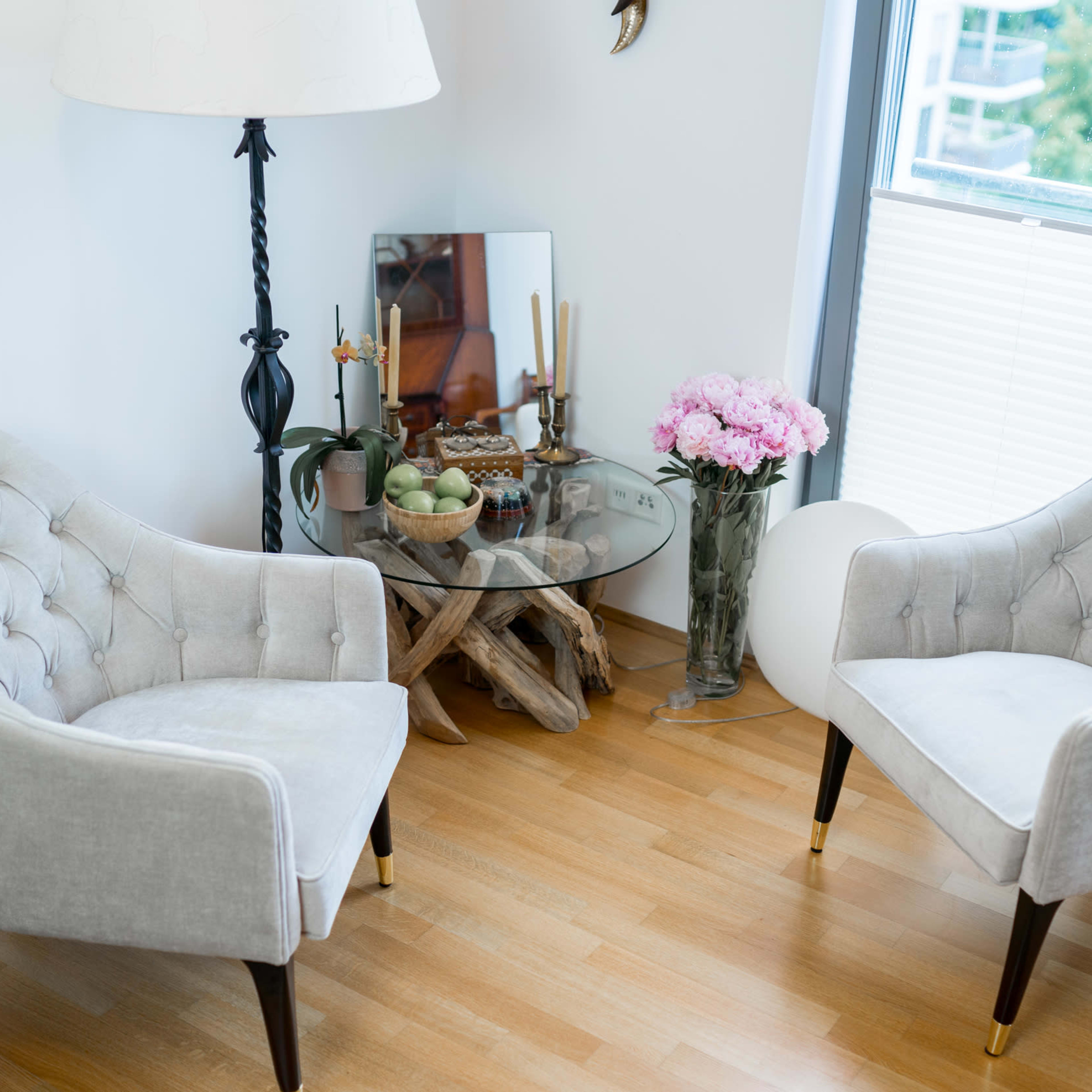A cozy seating area with two tufted chairs, a glass table on a wooden stand, a small vase of pink flowers, and various decorative items.