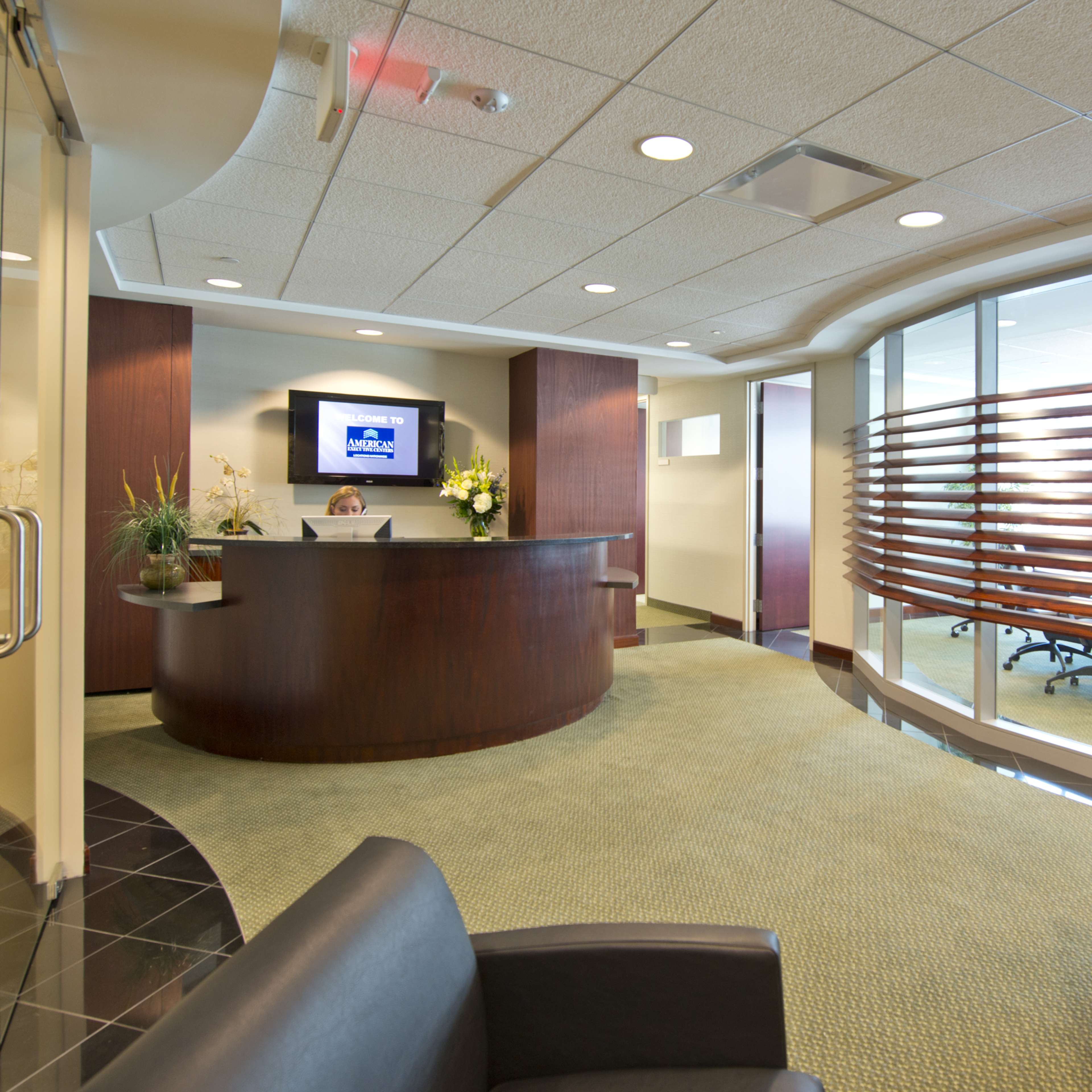 A modern office reception area with a curved reception desk, seating in the foreground, and a glass partition separating meeting spaces.