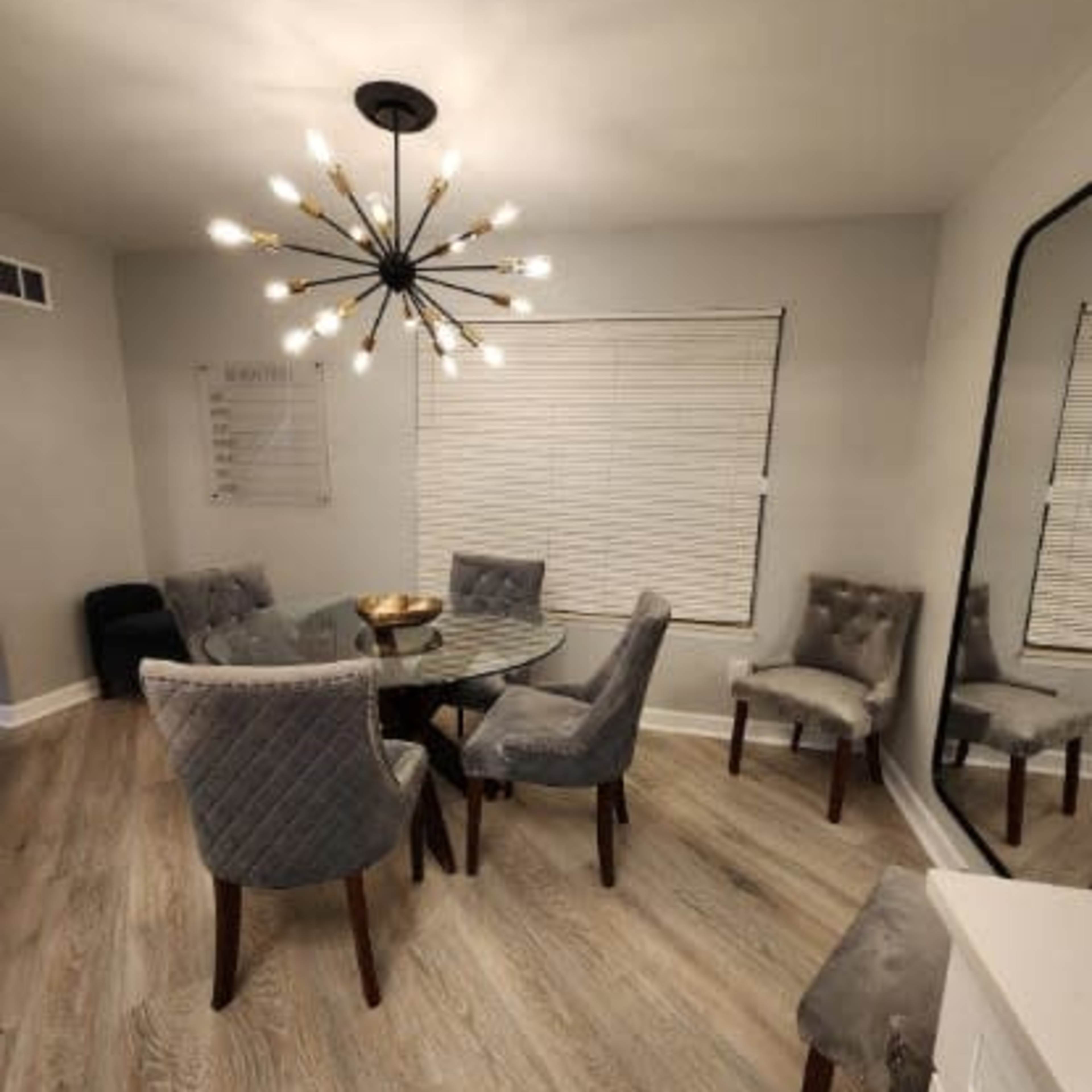 A modern dining area featuring a glass table surrounded by six upholstered chairs, with a large chandelier overhead and a mirrored wall.