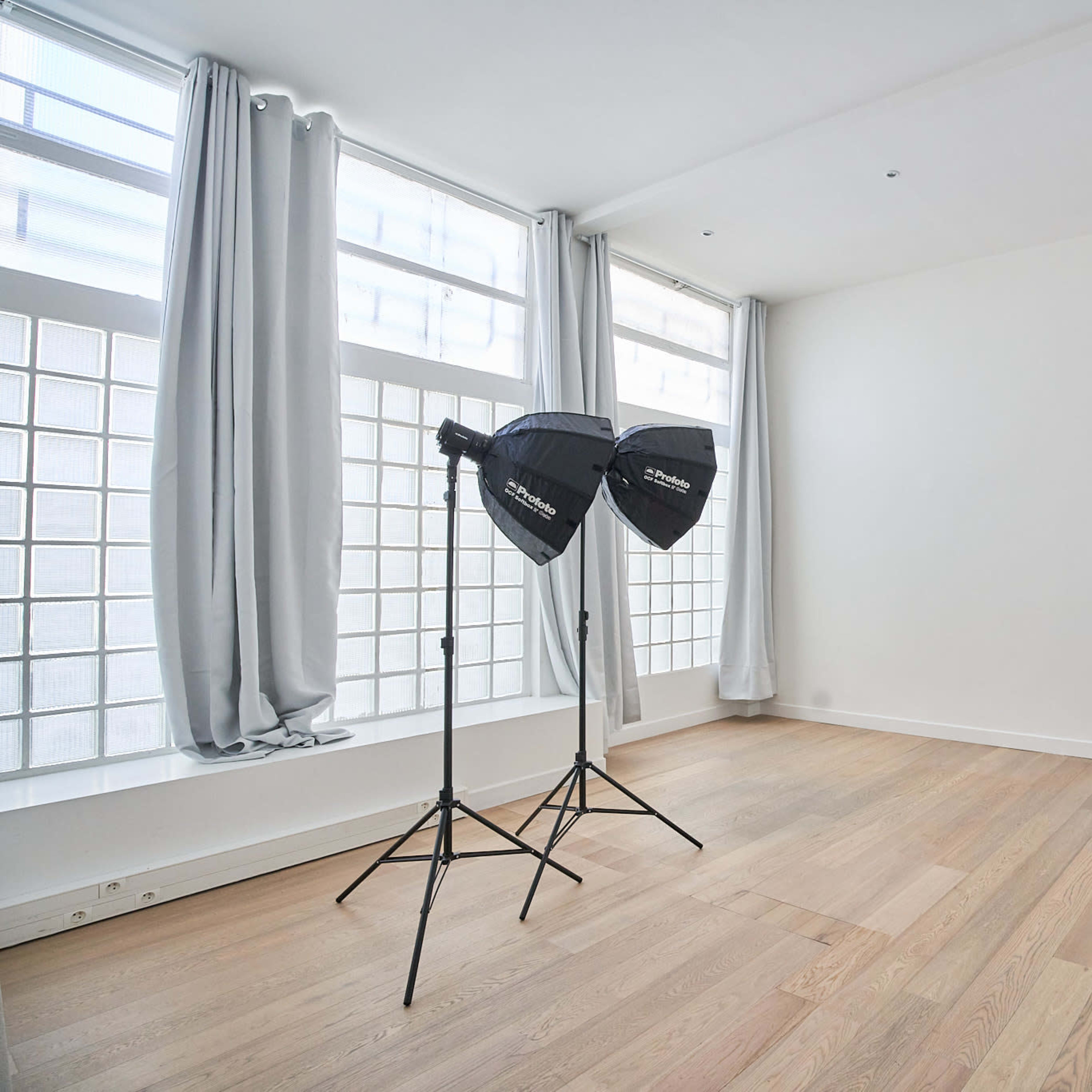A bright, empty room with large windows and two lighting softboxes on stands.