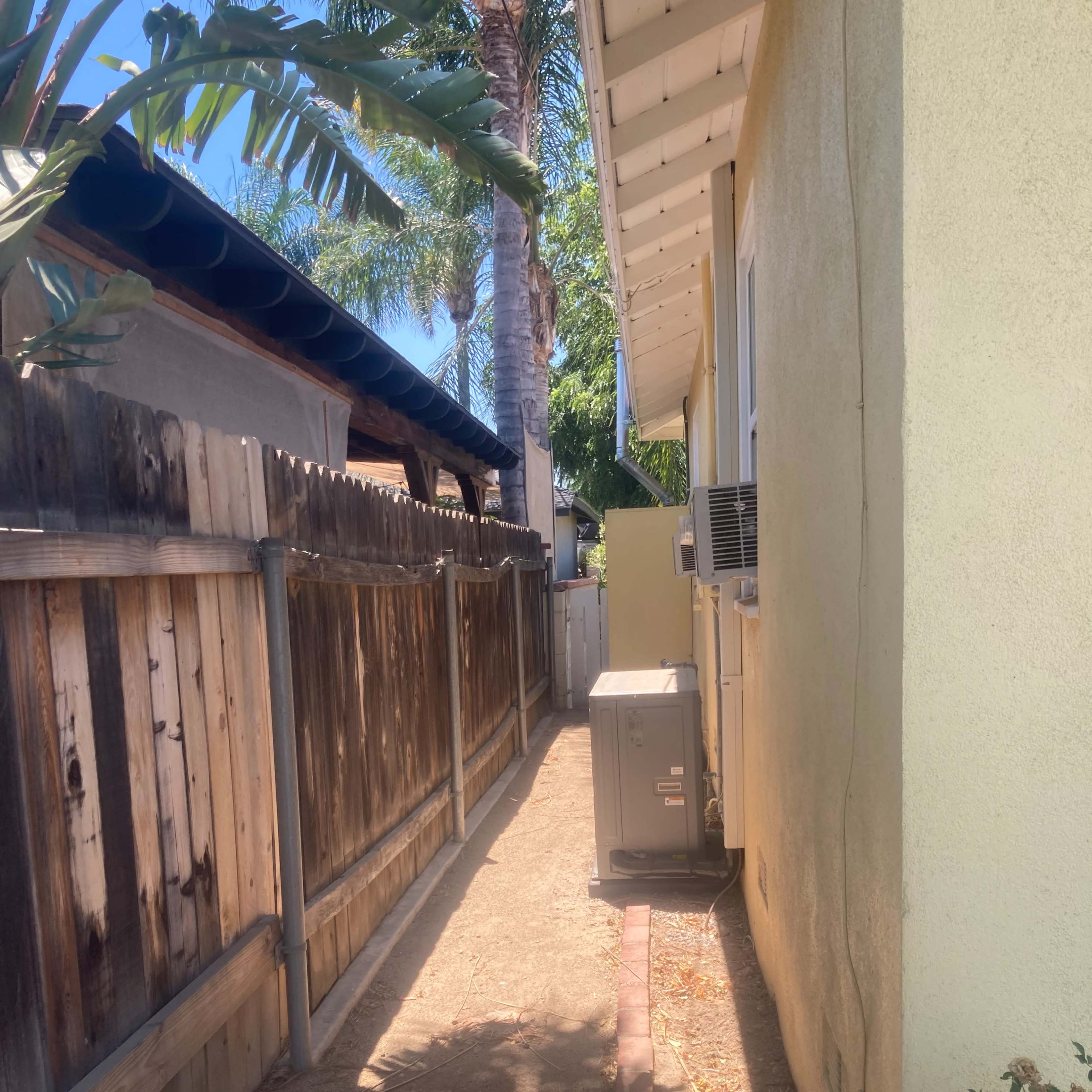 A narrow pathway runs between two houses, lined with a wooden fence and featuring an air conditioning unit along the yellow wall.