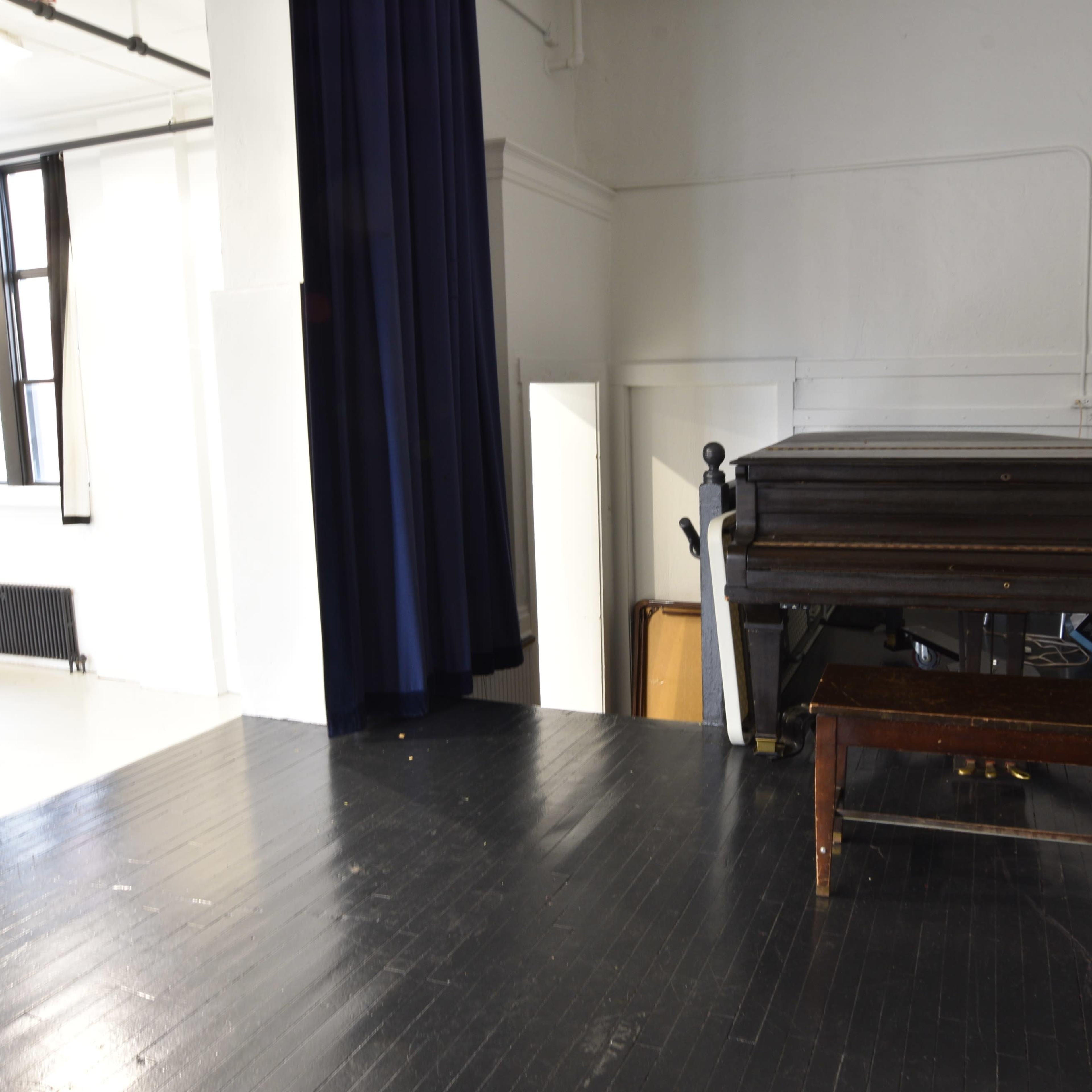 A stage area with a dark wooden piano and a matching bench on a polished black floor, surrounded by empty chairs and a curtain.