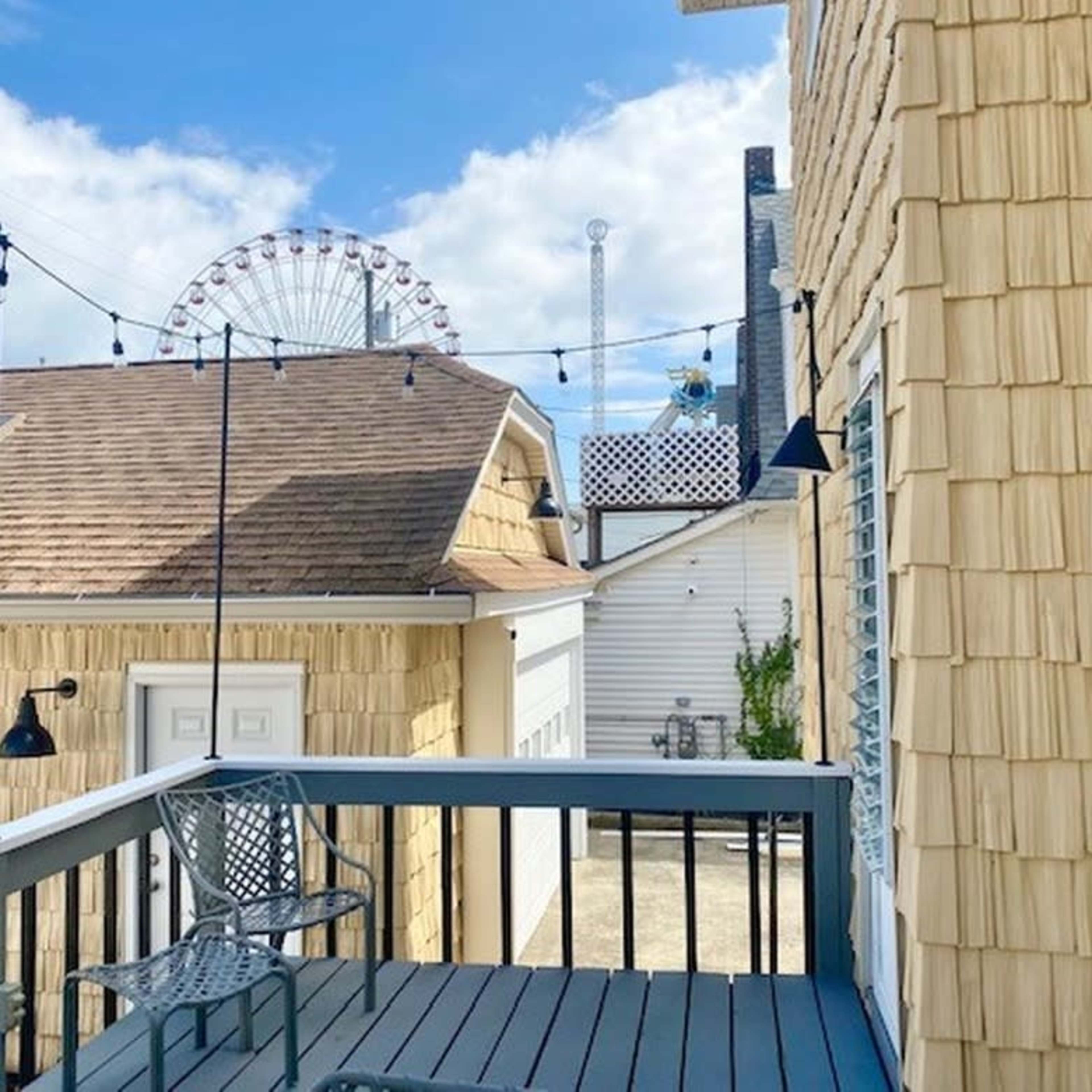 A balcony with a wooden deck and a chair overlooks buildings and a ferris wheel in the background under a partly cloudy sky.