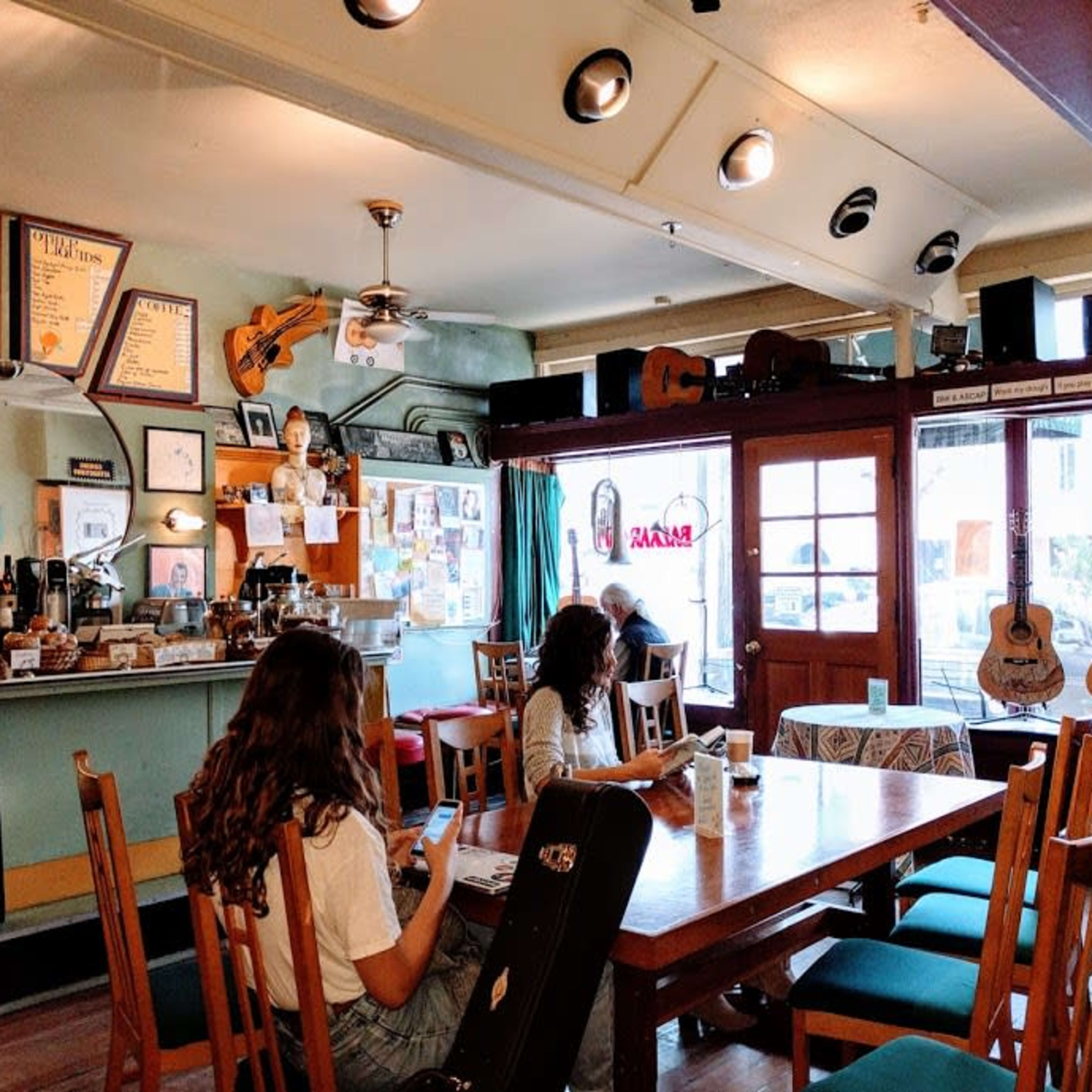 A cozy cafe features a wooden table surrounded by patrons, with walls adorned with musical instruments and framed photographs.