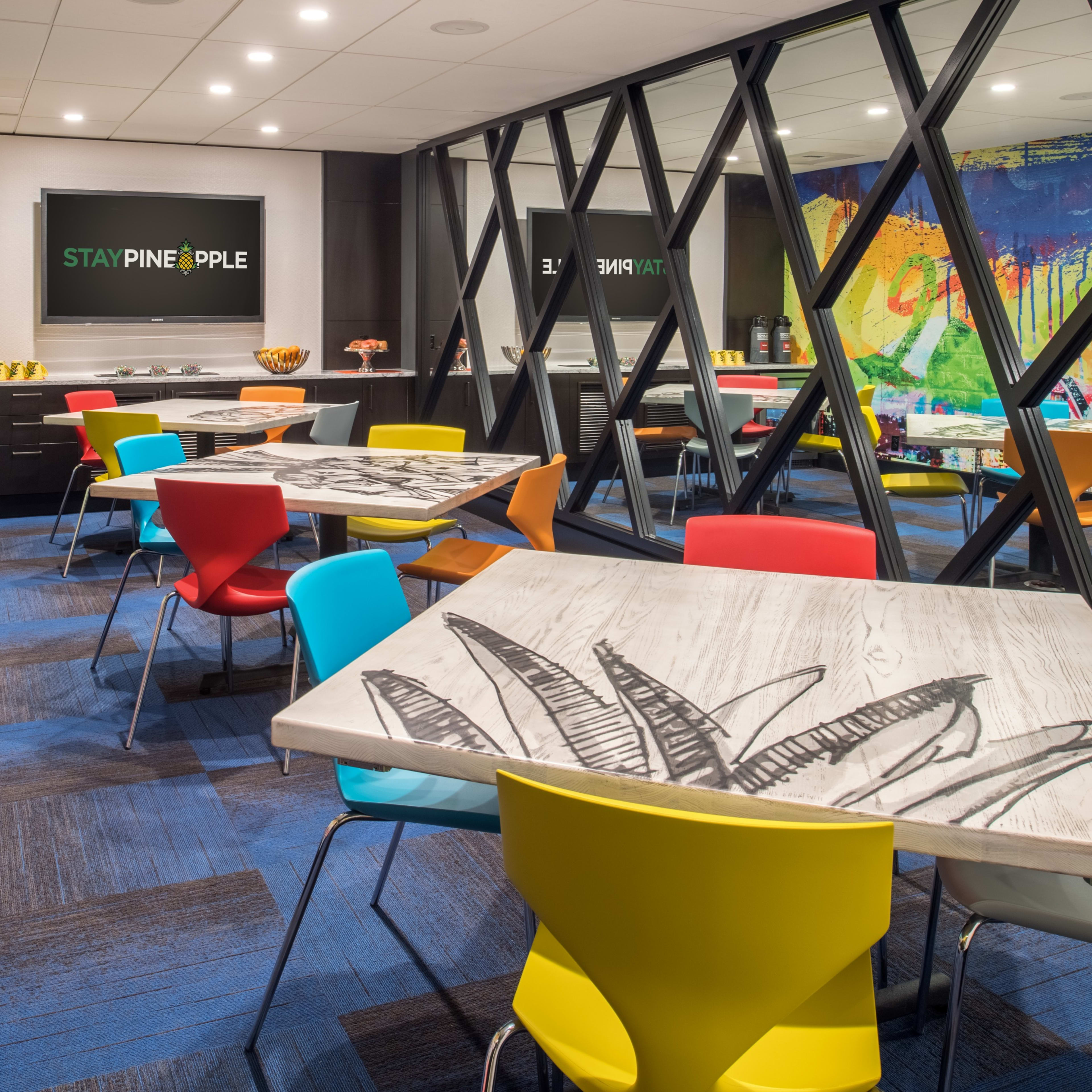 A colorful break room with tables and chairs arranged around a central area, featuring large windows and an artistic mural on the walls.