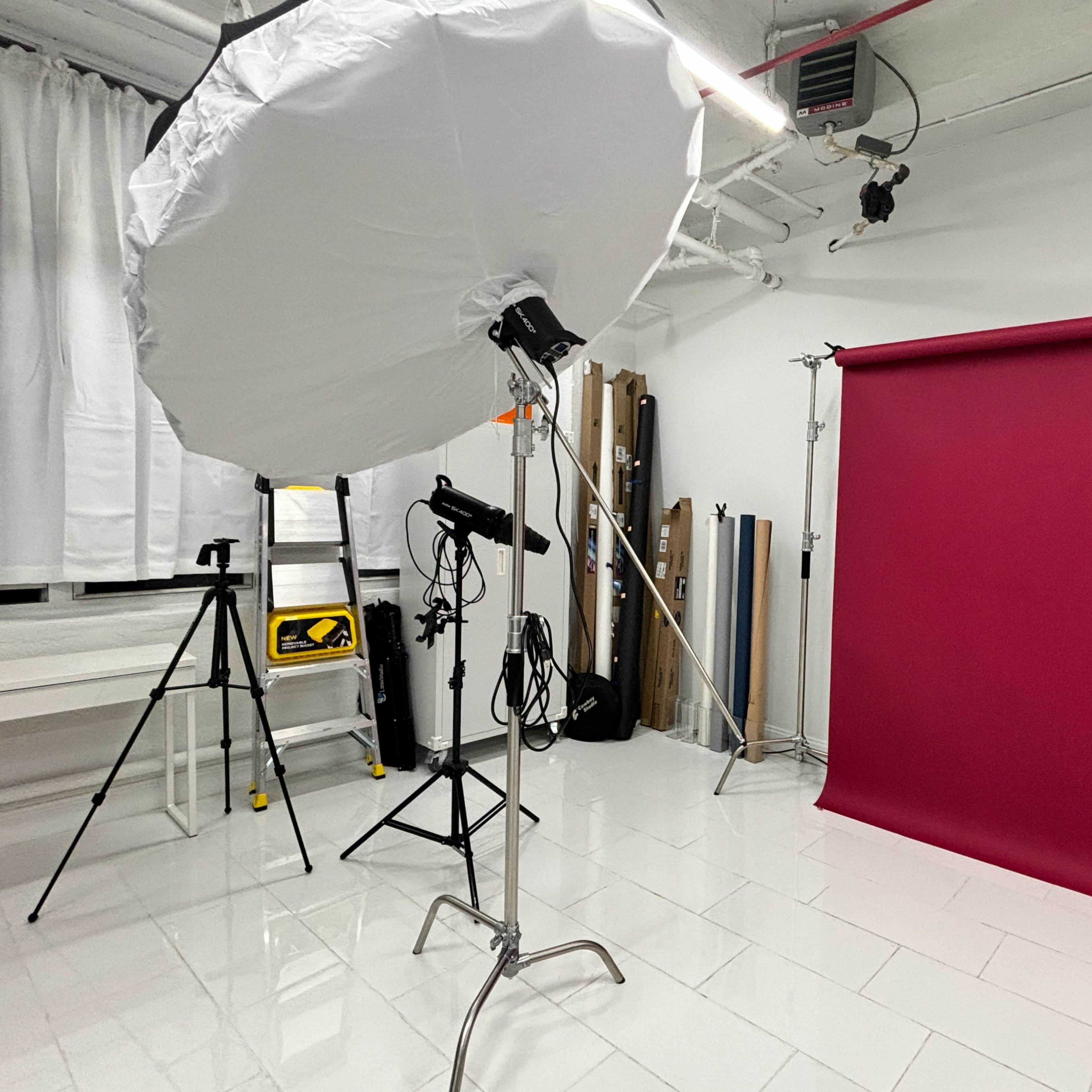 A photography studio setup with a large softbox light, a tripod, and a solid burgundy backdrop against a white tiled floor.