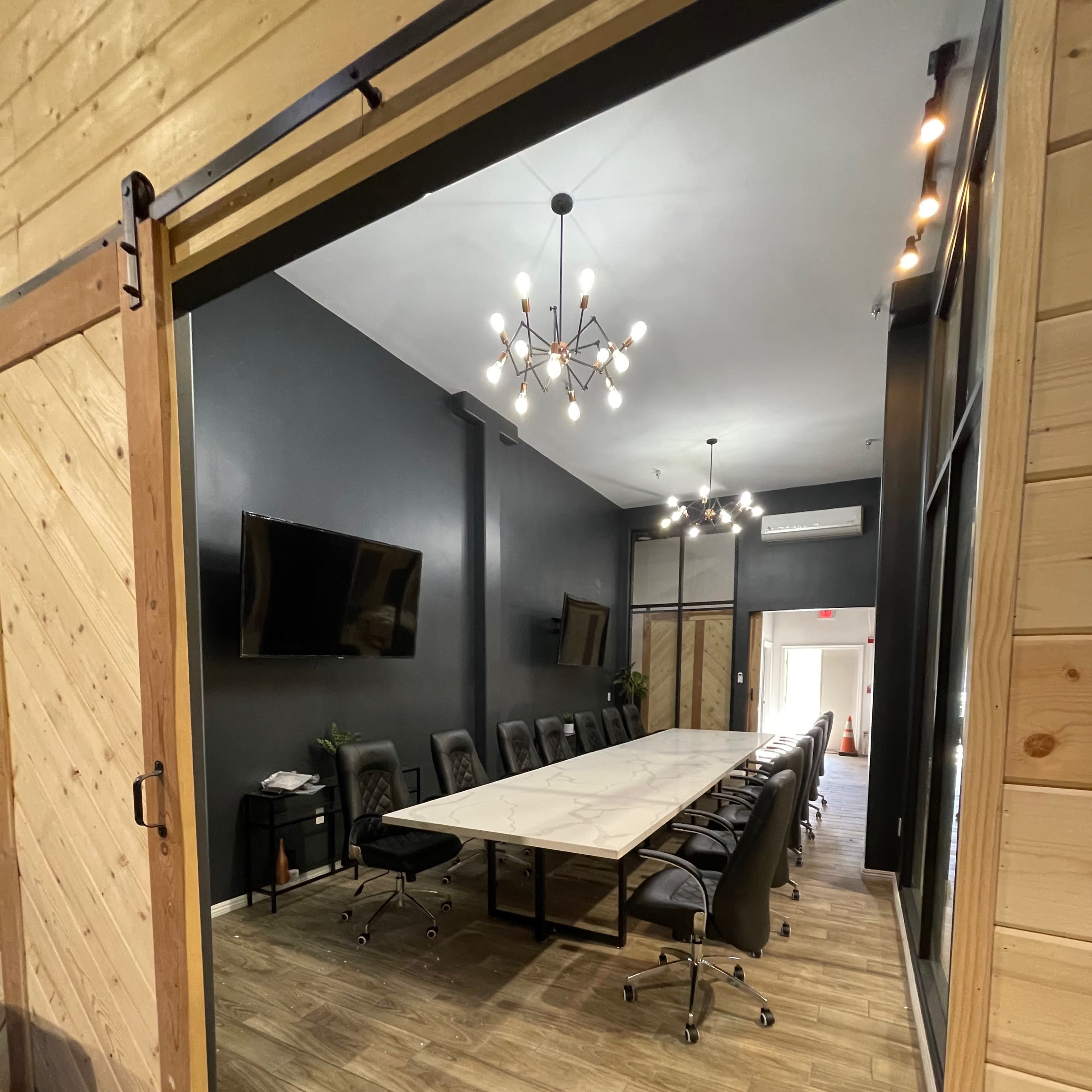 A modern conference room with a long table, black chairs, and large screens mounted on the dark walls.