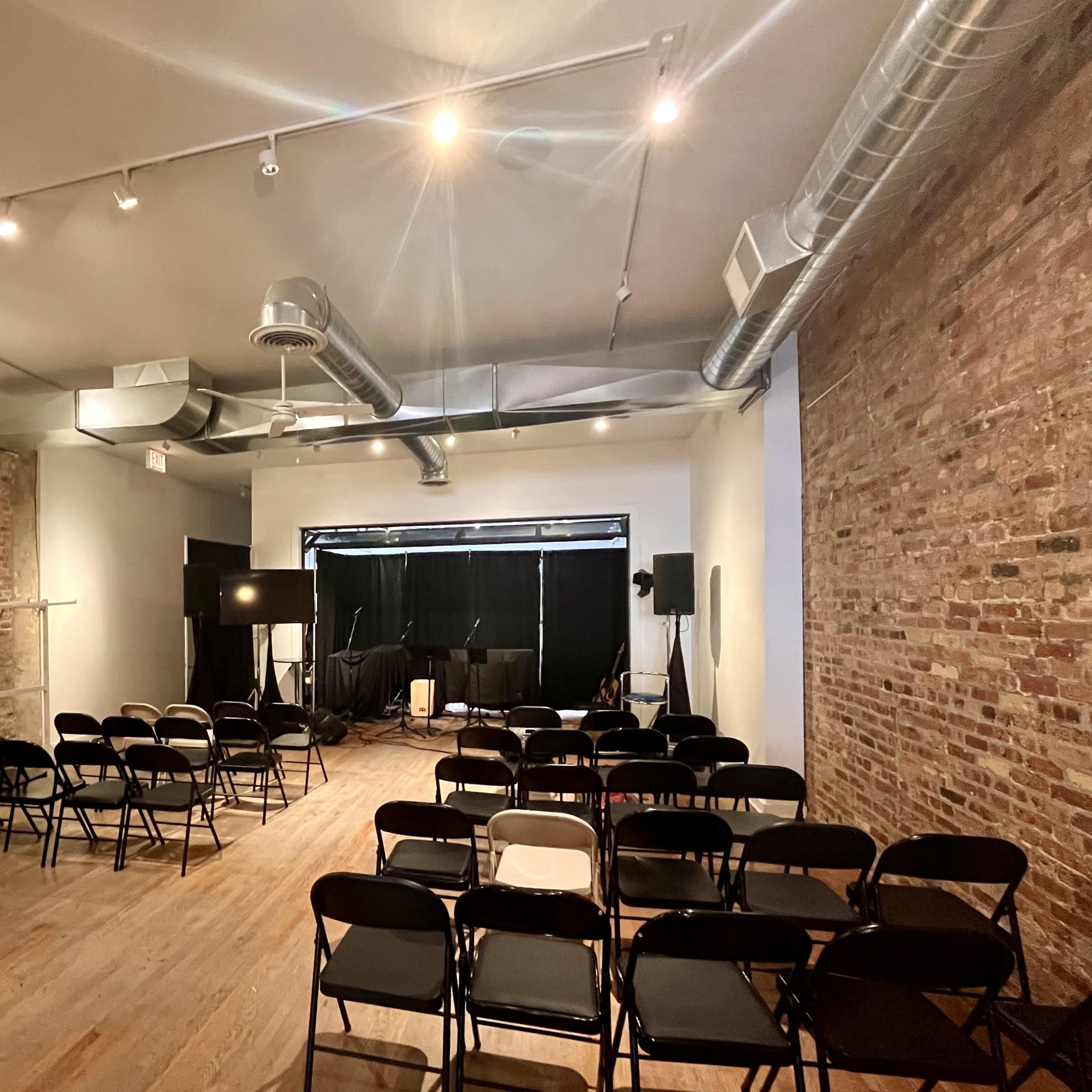 A minimally furnished event space with wooden flooring, exposed brick walls, and rows of black chairs set up for an audience facing a stage area.